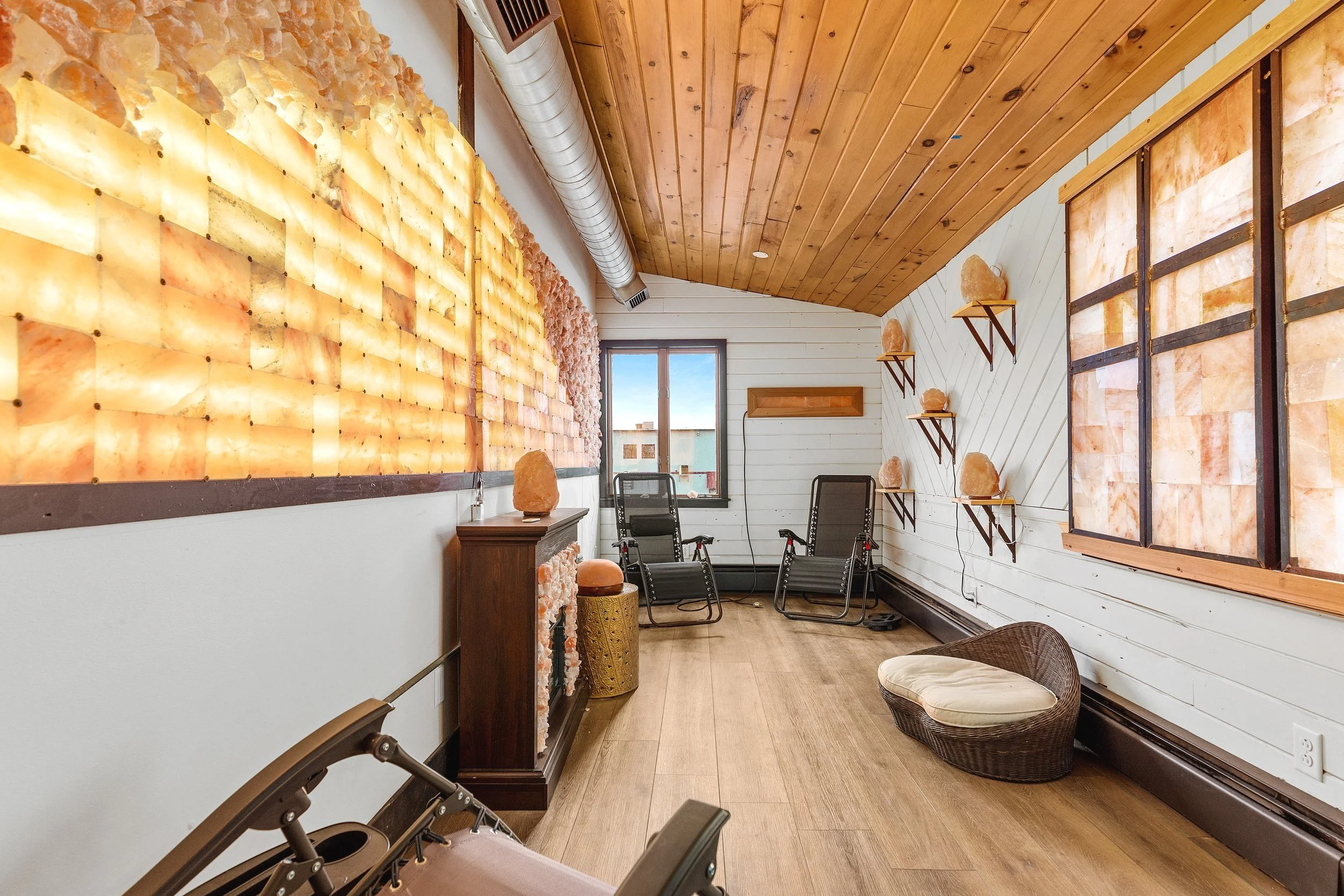 A cozy room with salt lamps on the walls and shelves, two black chairs near the window, a wicker pet bed, and a salt lamp on a wooden pedestal.