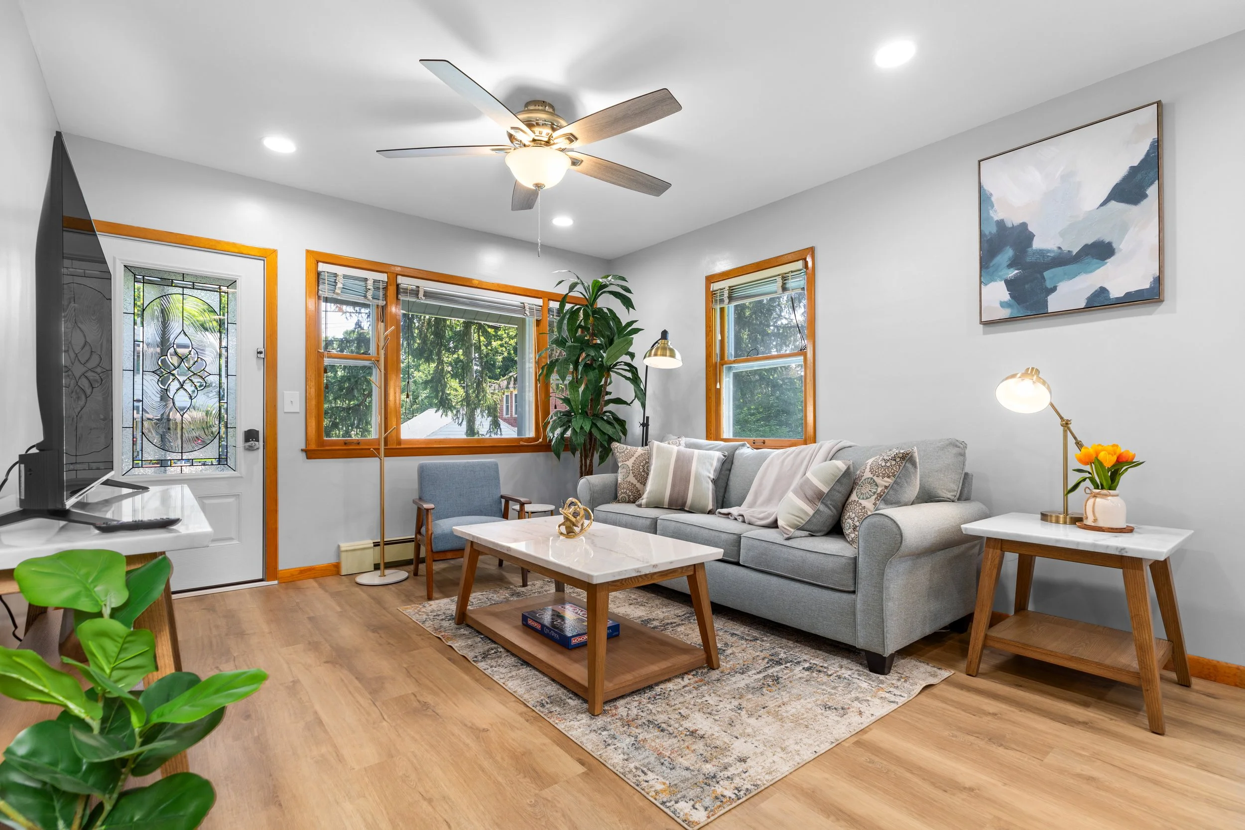 Bright living room with gray sofa, wooden coffee table, side table with lamp and flowers, large windows with wooden trim, ceiling fan, artwork on wall, and hardwood floors.