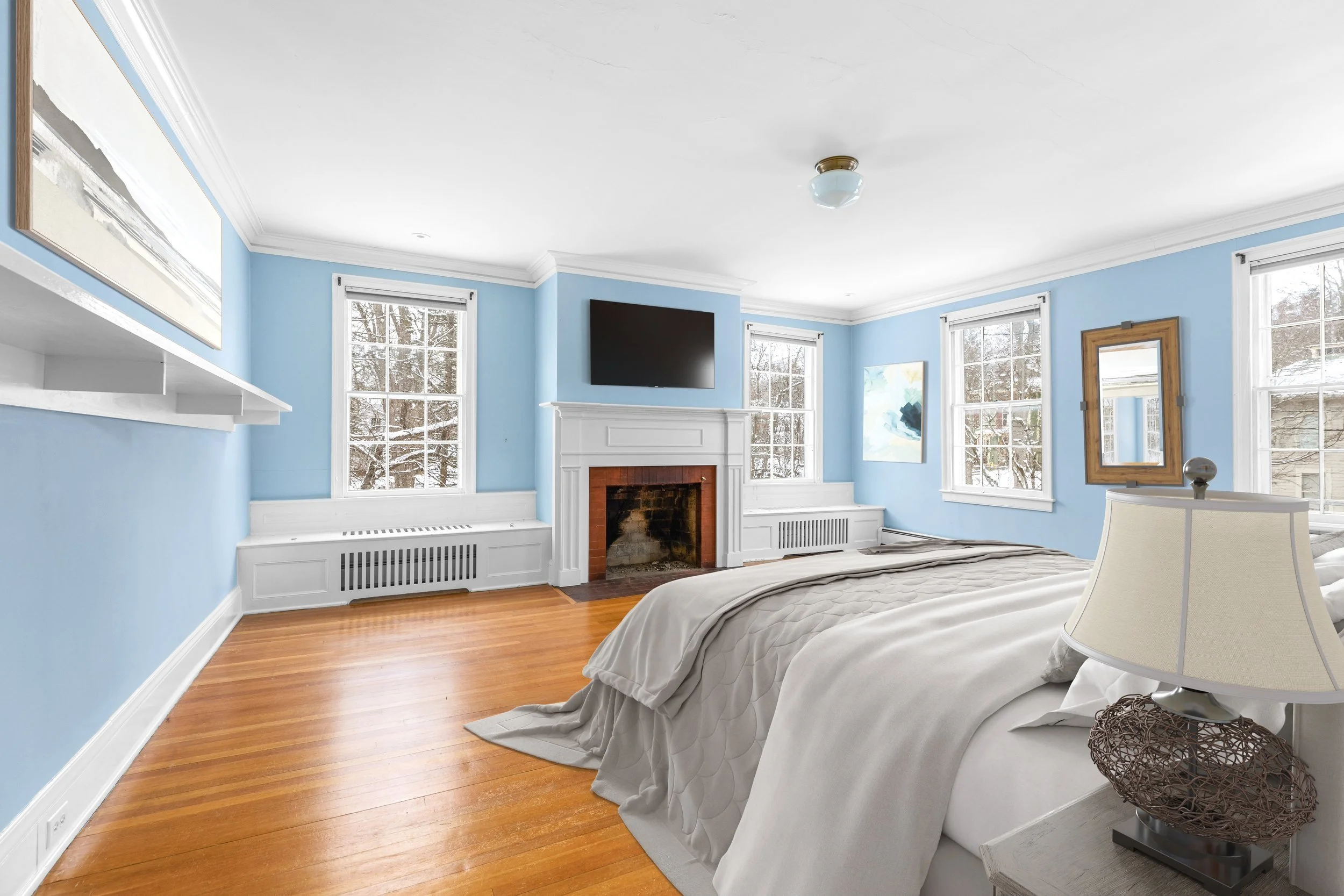 A bright bedroom with light blue walls, multiple windows, a wooden floor, and a white bed with beige bedding. There is a white fireplace with a black screen above it, a flat-screen TV mounted on the wall, and framed art. A nightstand with a lamp is b