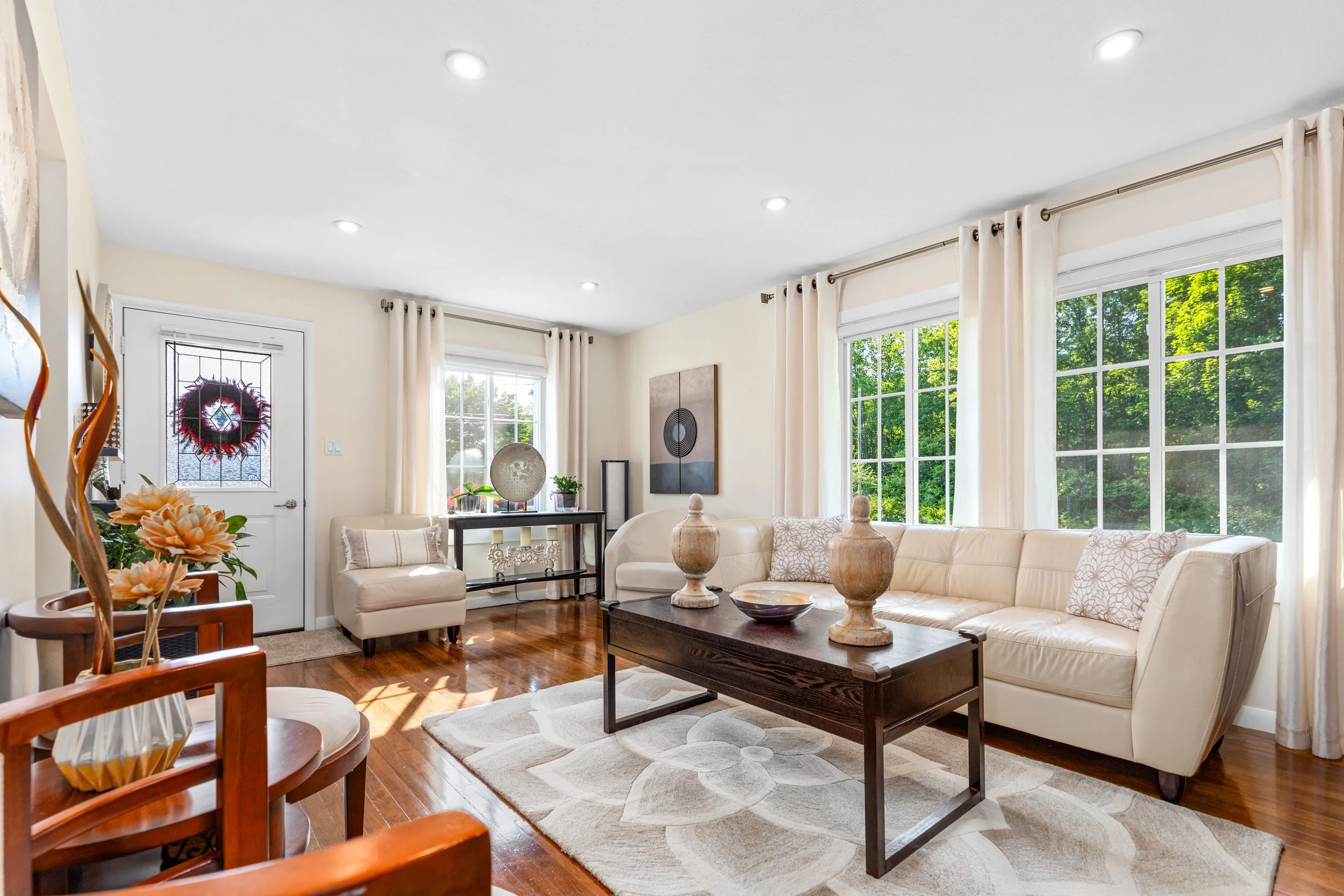 Bright living room with large windows, cream sofa, dark wood coffee table, decorative vases, artwork, and hardwood flooring.
