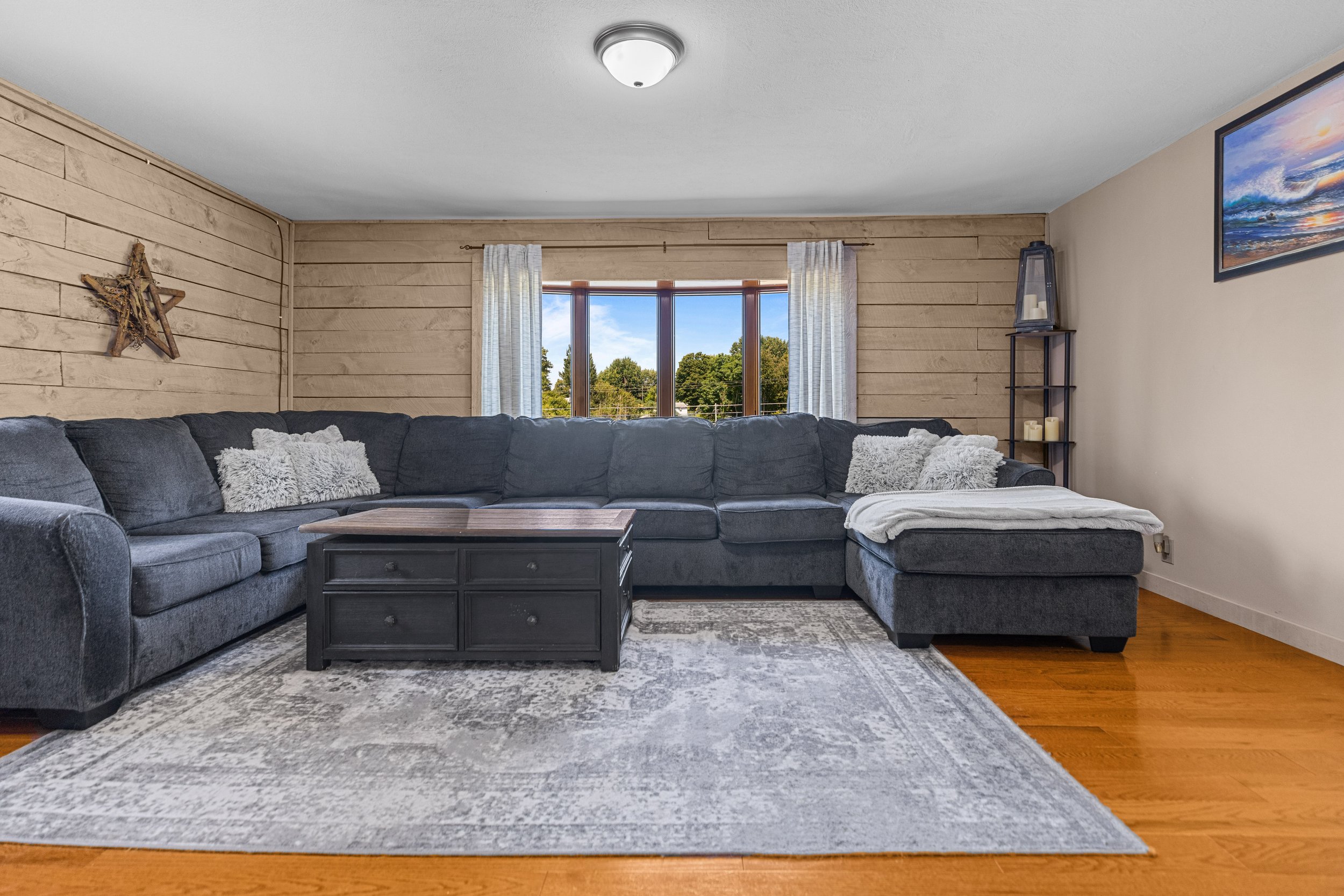 Living room with a large dark gray sectional sofa, light-colored curtains, a wooden coffee table, a light-colored rug, a window with trees outside, a wall with a decorative star, and a framed landscape photo.