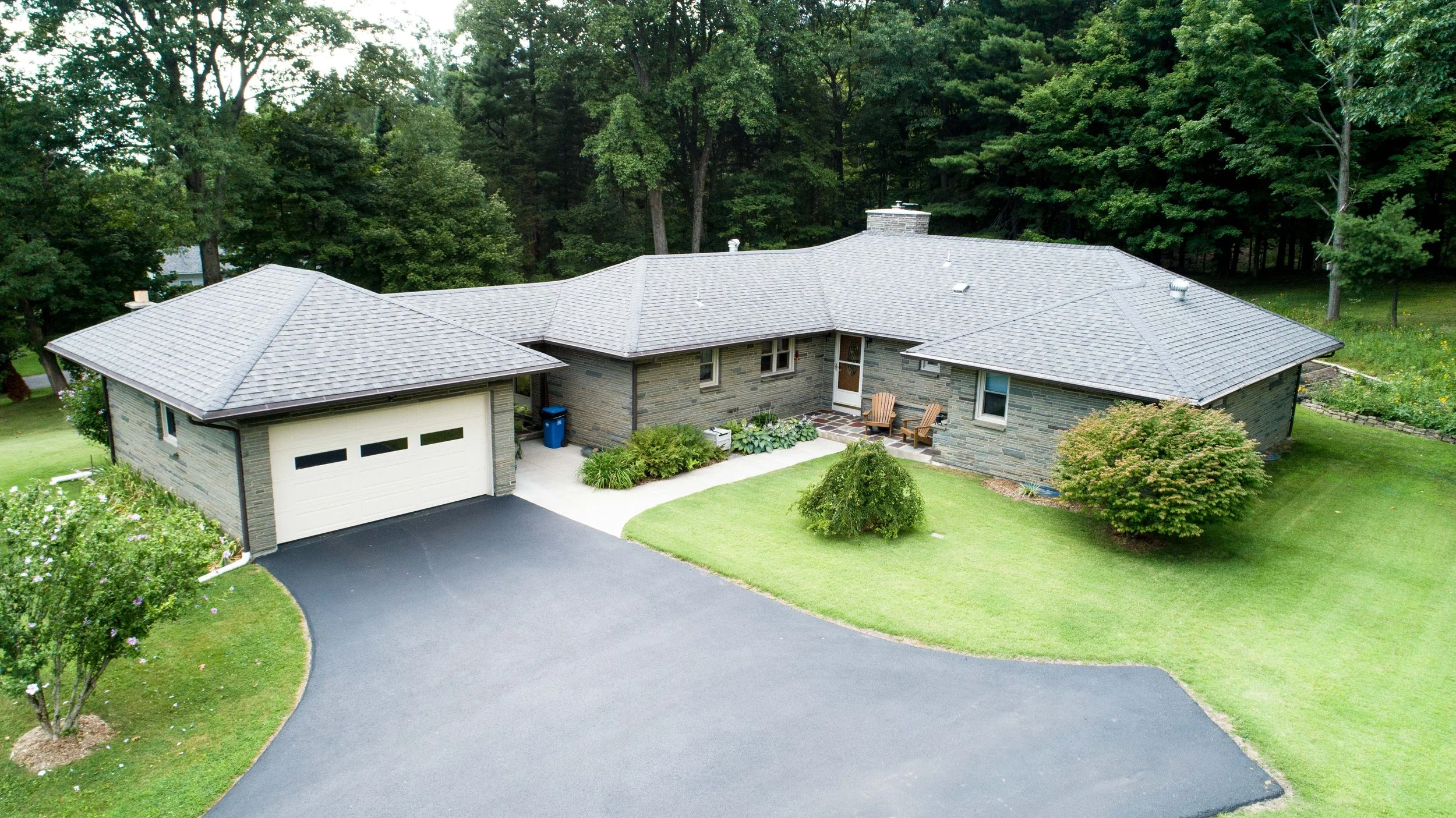 A single-story house with a gray shingle roof, brick exterior, attached garage, front yard with green grass, trees, and shrubs, and a driveway leading up to the garage in a wooded area.