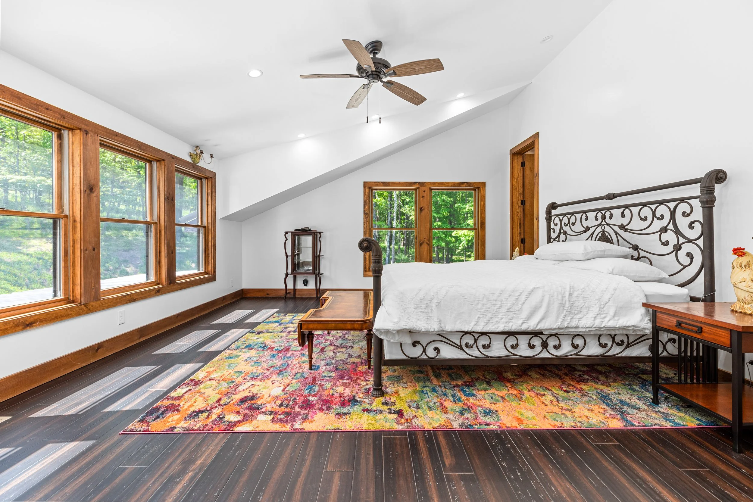 A bedroom with large windows showing green trees outside, a white bed with a decorative metal headboard, a colorful area rug, wooden furniture, and a ceiling fan.