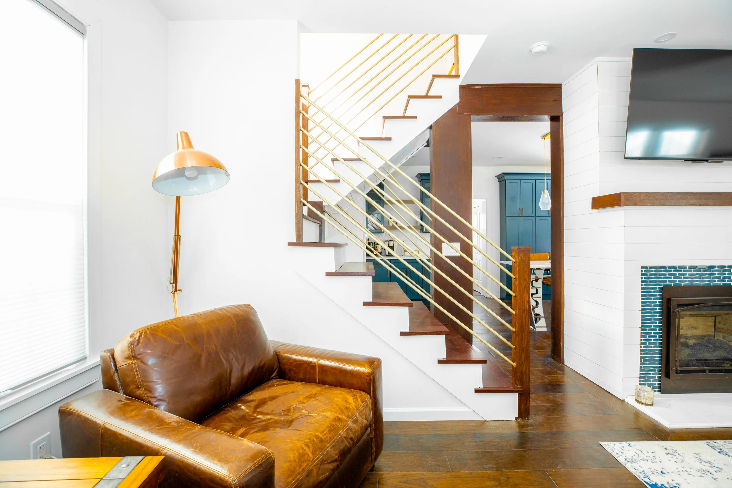 Living room with a brown leather sofa, a tall gold and copper floor lamp, a staircase with wooden steps and gold railing, a white shiplap fireplace with blue tile, a mounted flat-screen TV, and a glimpse of a kitchen with blue cabinetry and a dining 