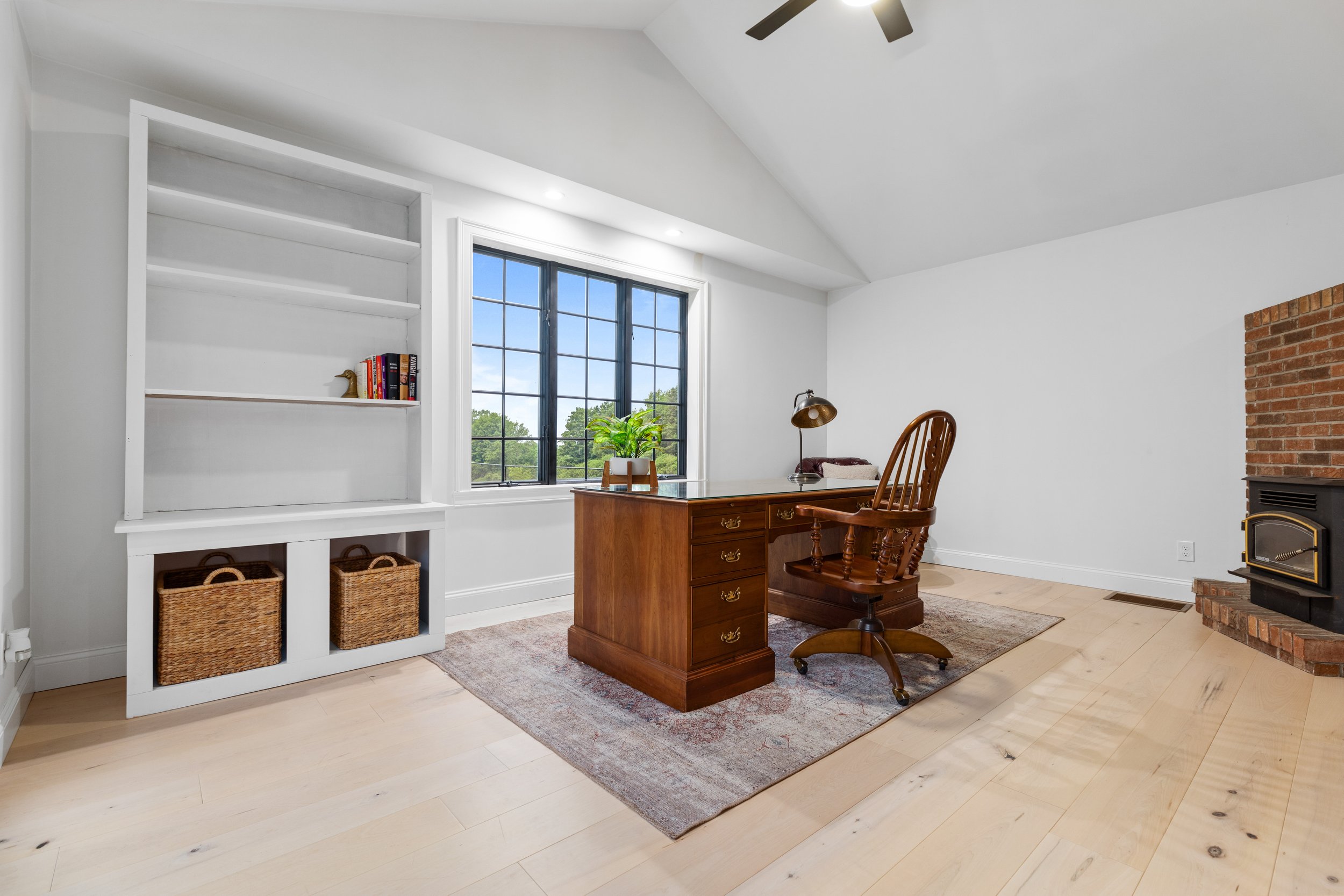 Home office with wooden desk and chair, bookshelf, window with blue sky, brick fireplace, ceiling fan, light-colored hardwood floors, area rug, potted plant, lamp.