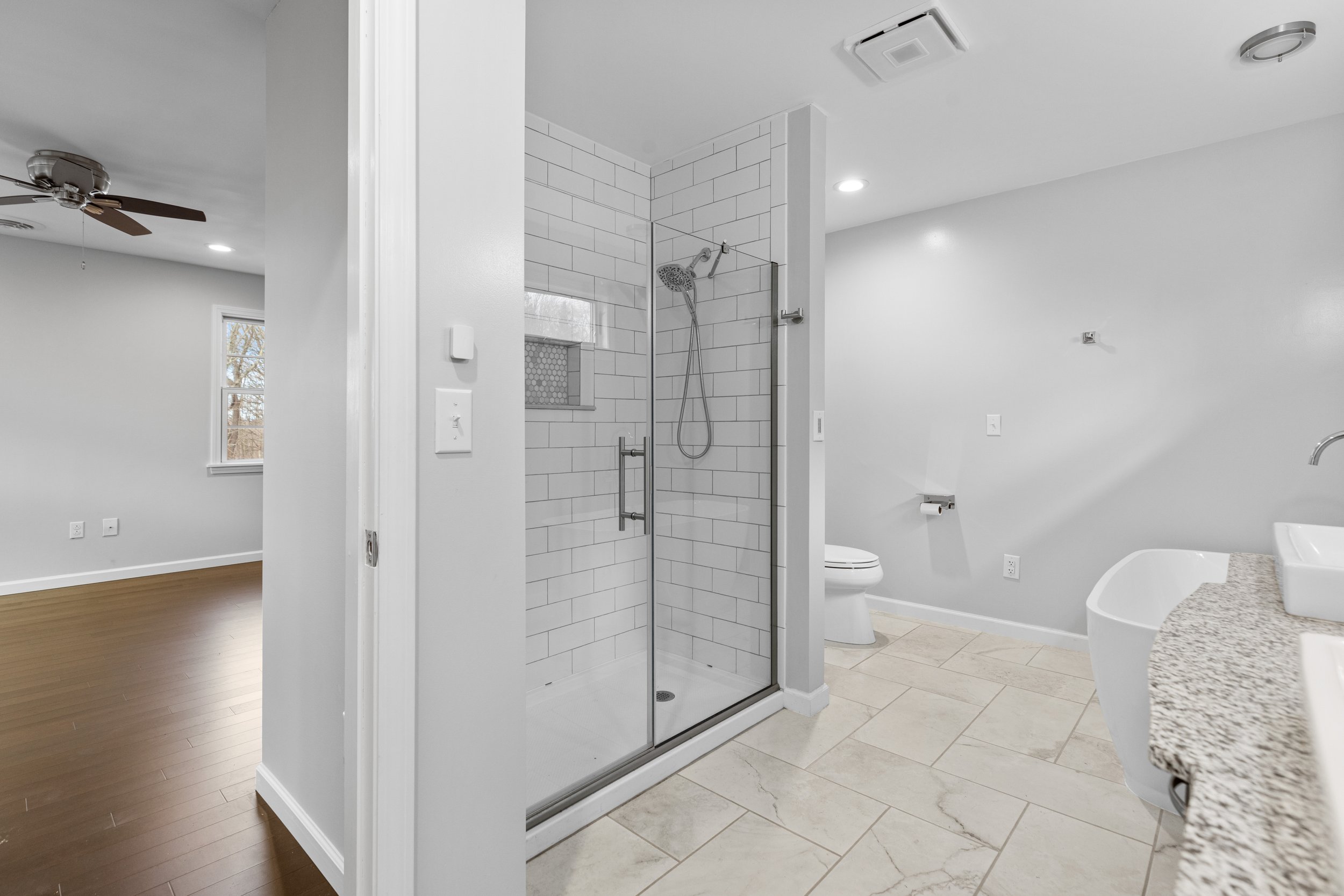 Bathroom with a shower, toilet, and countertop with a sink, adjacent to a room with hardwood floors and a ceiling fan.