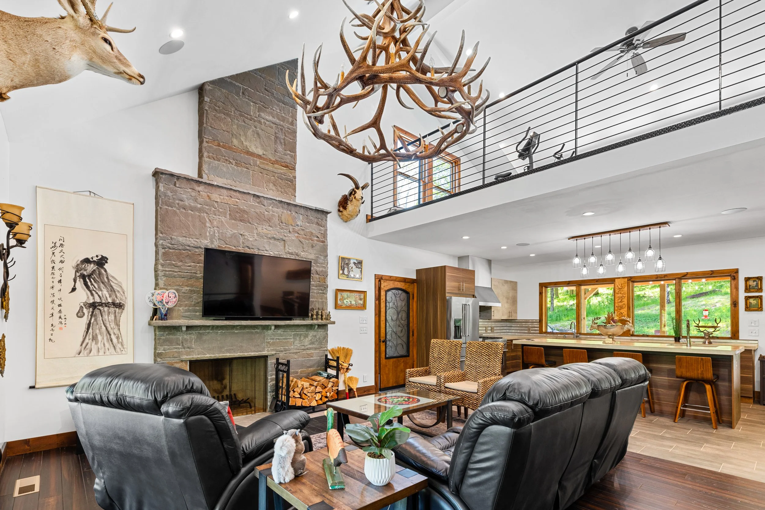 Interior of a living room with a stone fireplace, mounted television, recliner chairs, a coffee table, and various decorative mounts of animals including a deer head and antlers on the ceiling. The room has a high ceiling, large windows, and an open 