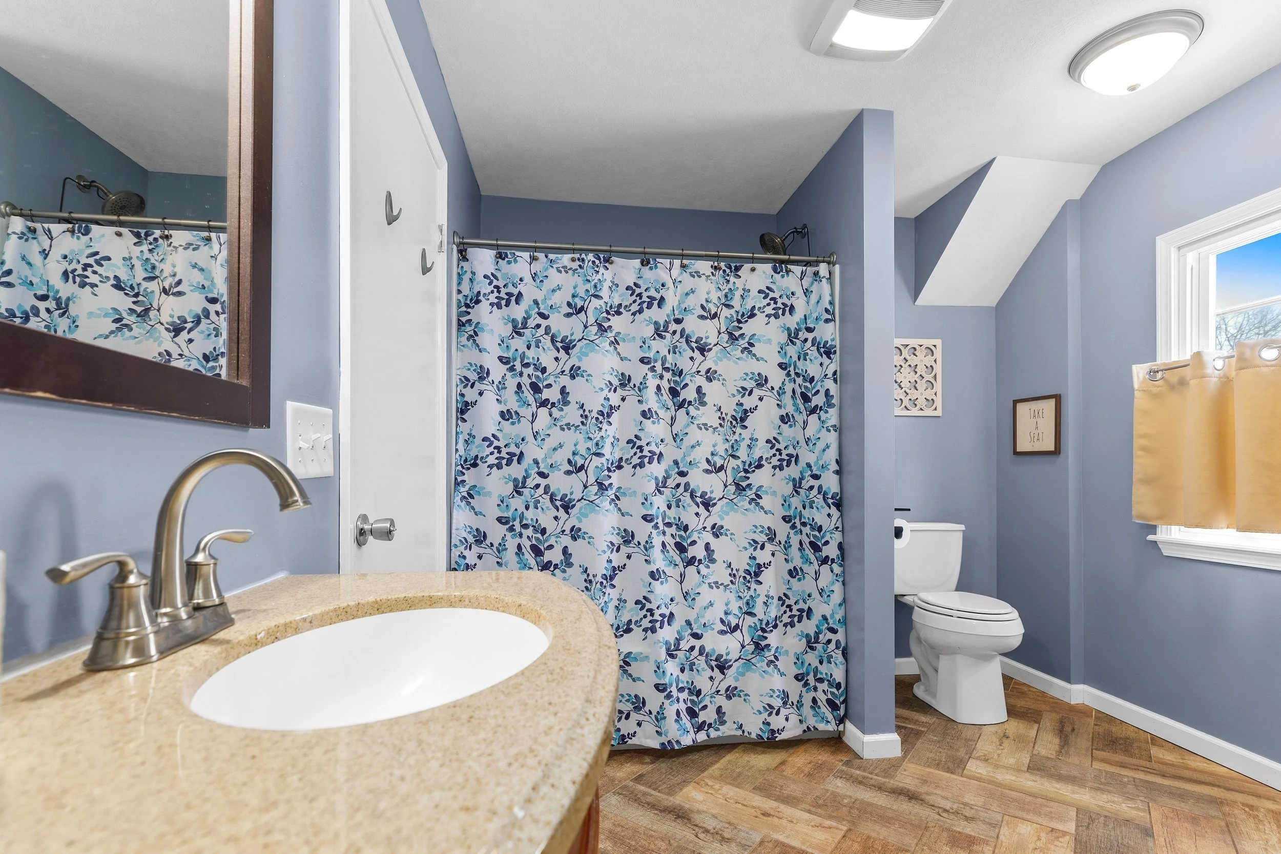 A bathroom with a blue wall, a beige countertop with a sink, a mirror, a shower with a floral curtain, a toilet, and a window with beige curtains.