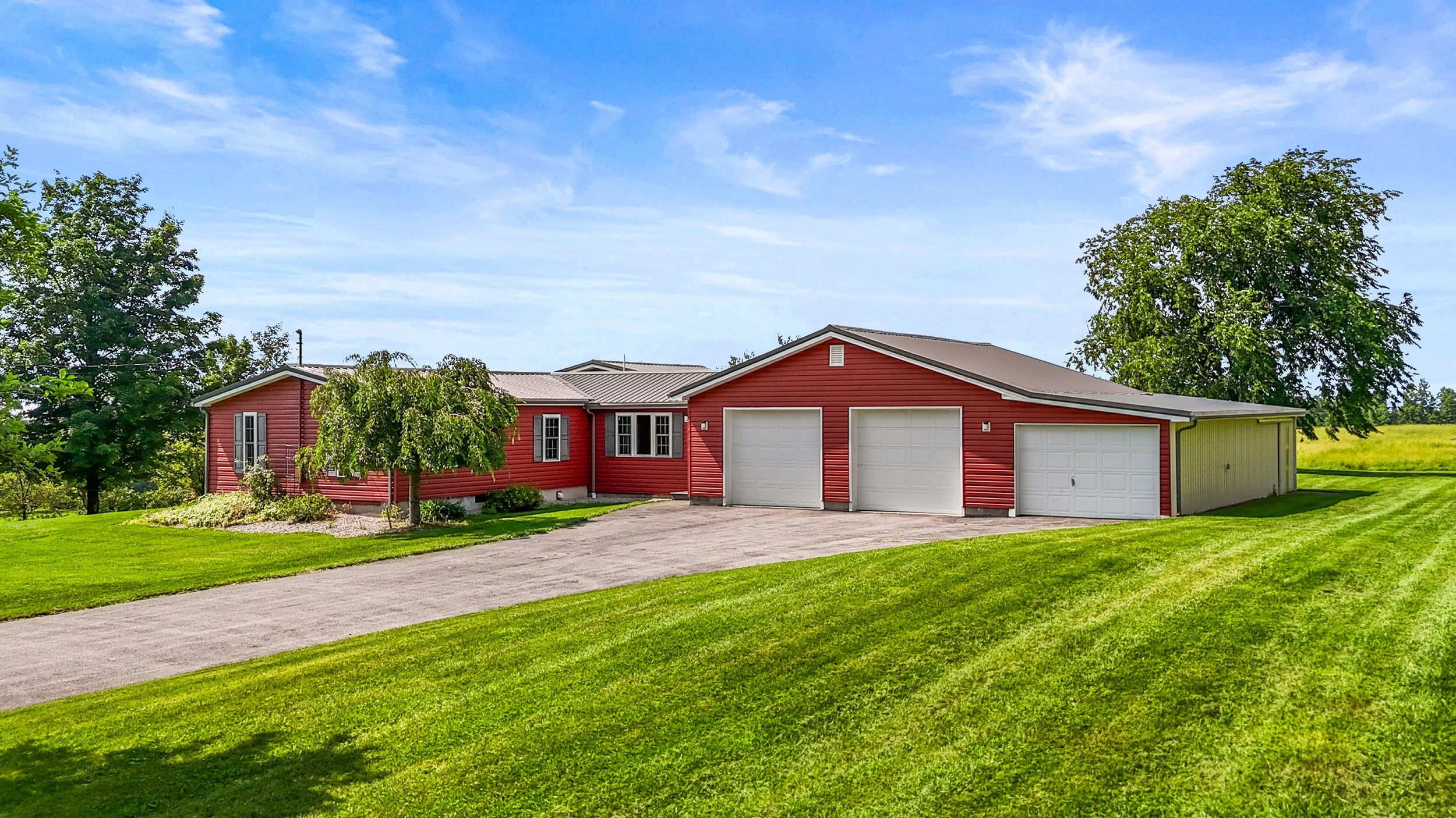 A red house with a three-car garage, surrounded by green grass and trees, under a partly cloudy sky.
