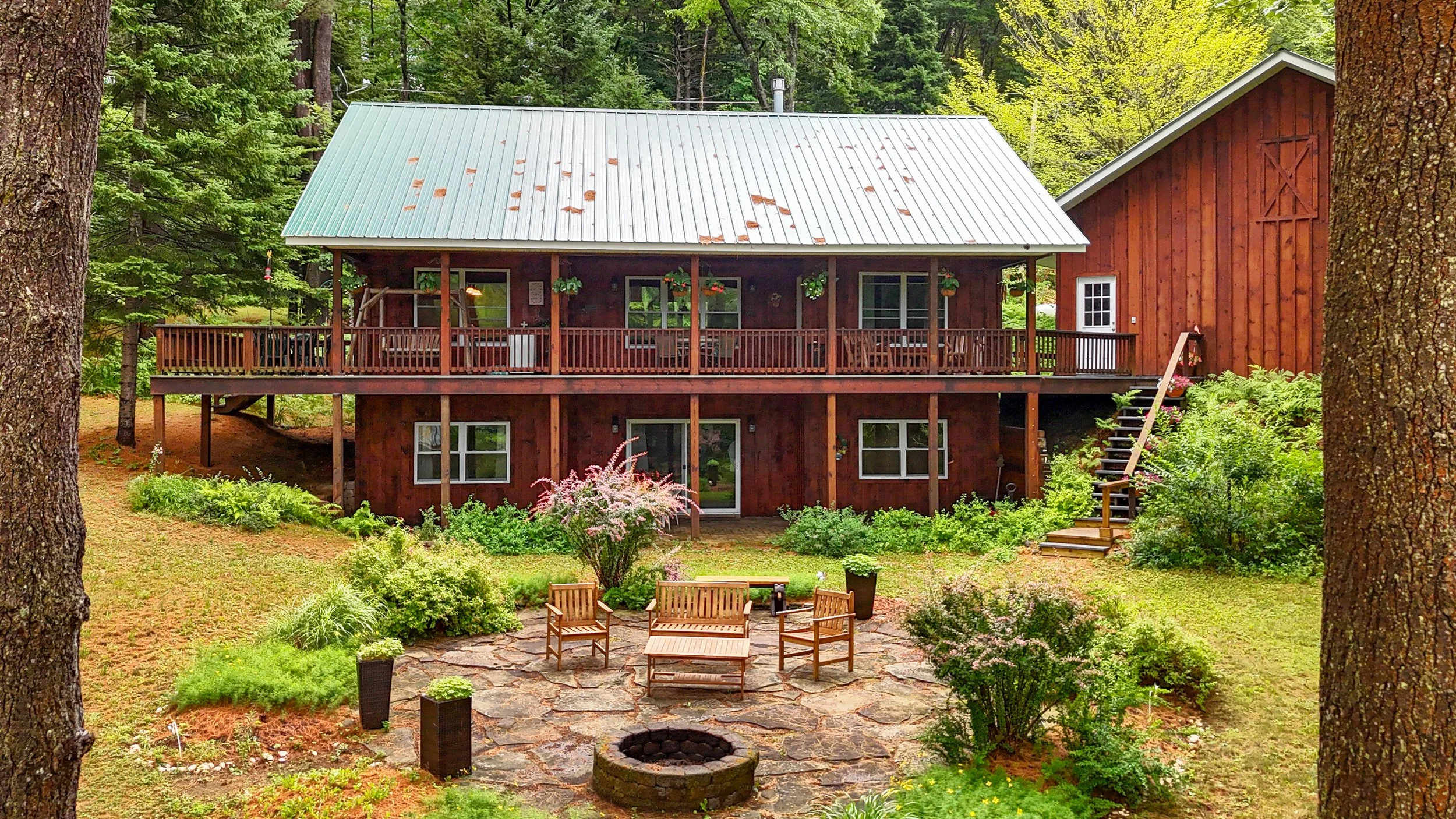 A rustic two-story cabin in the woods with a large screened porch and a deck, surrounded by trees and lush green landscaping, including a patio with outdoor furniture and a fire pit.