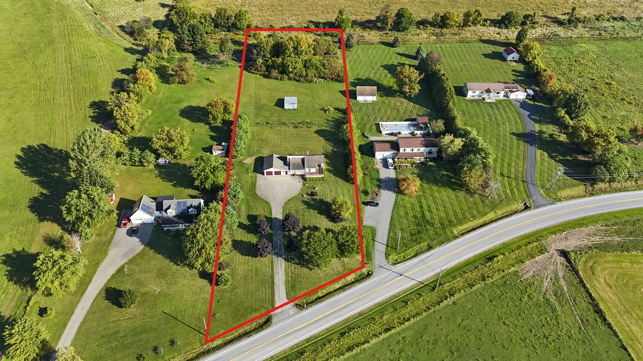 Aerial view of a large property outlined in red, featuring a house with a driveway, various trees, smaller outbuildings, and expansive green lawns, situated along a curved two-lane road.