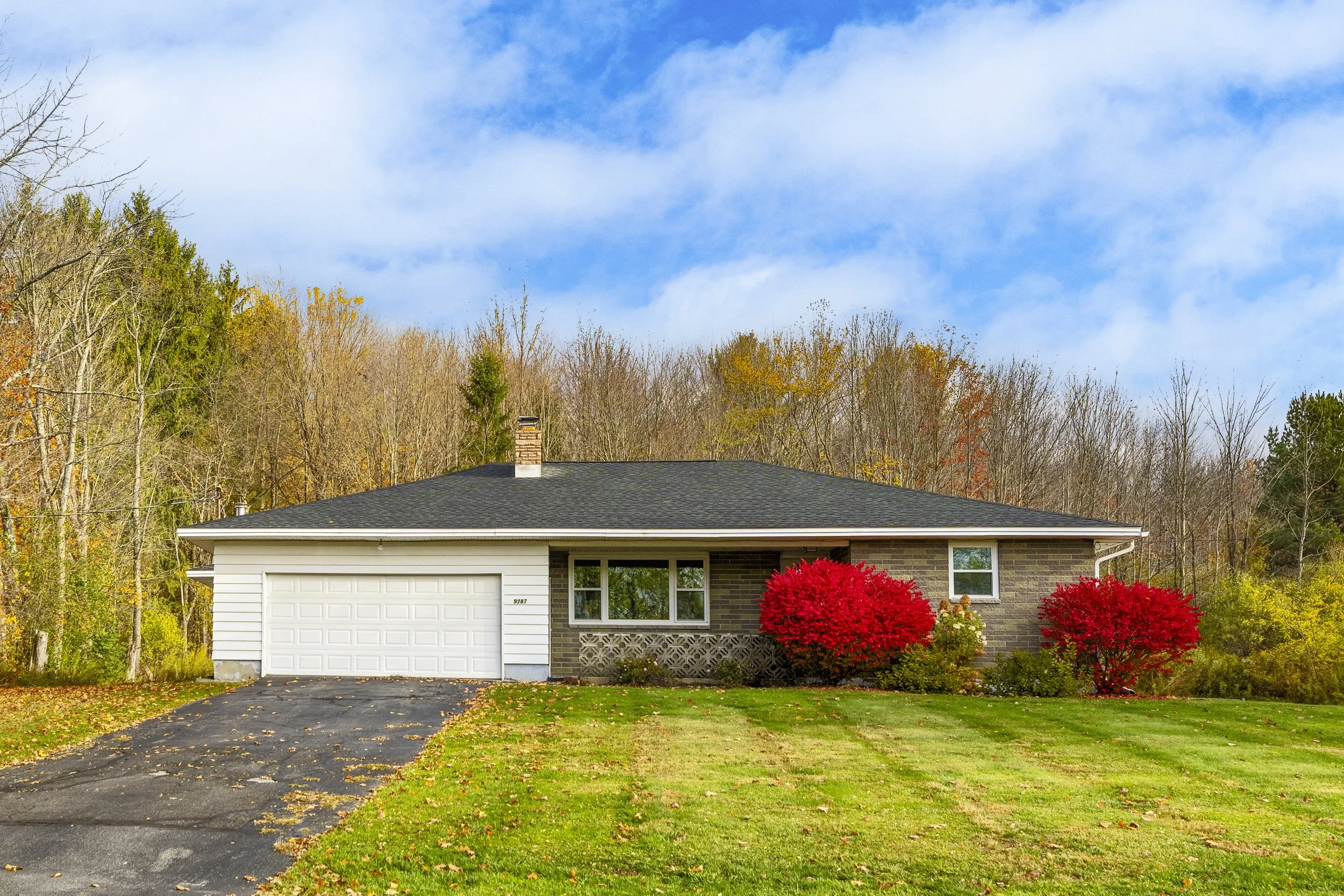 A single-story house with a black roof, white garage door, and brick exterior, surrounded by colorful autumn foliage and a well-maintained lawn.
