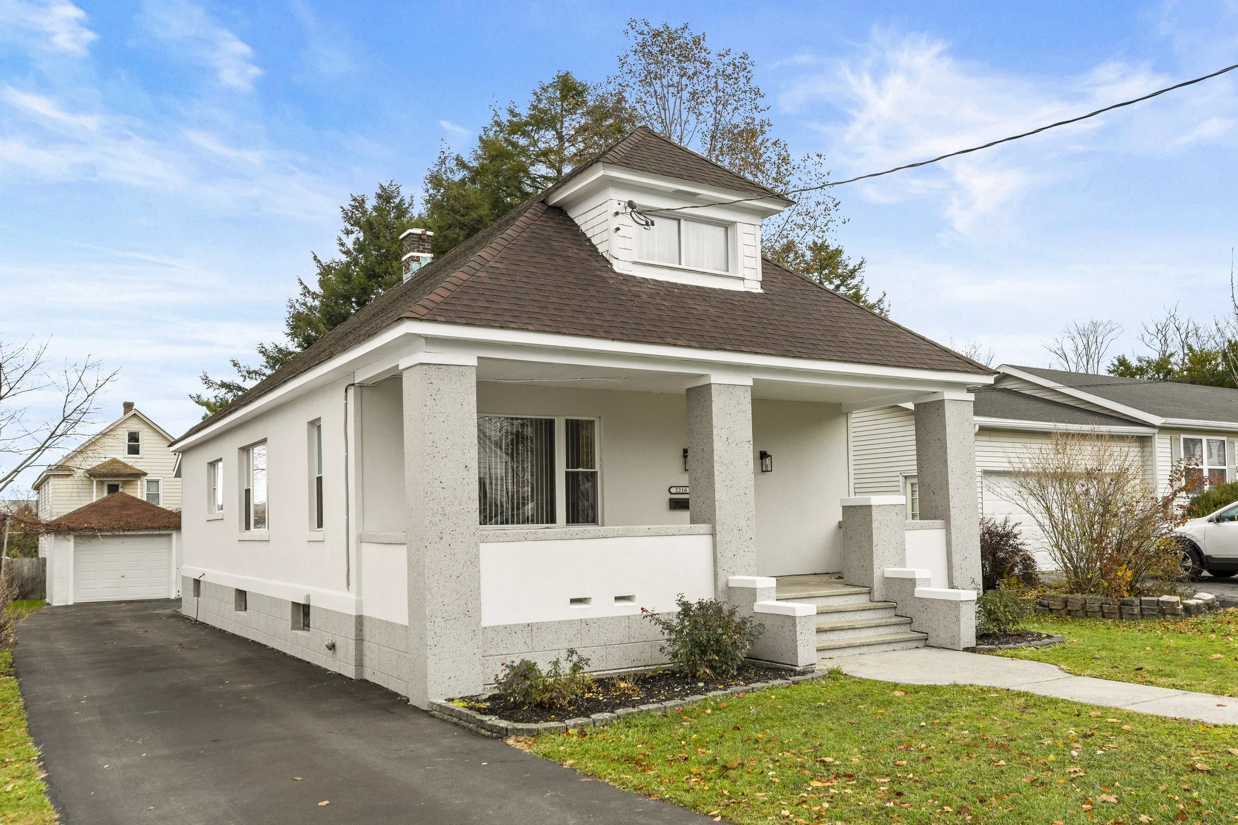 White house with a brown roof, front porch with steps, and small garden in front, situated in a neighborhood with other houses and trees.