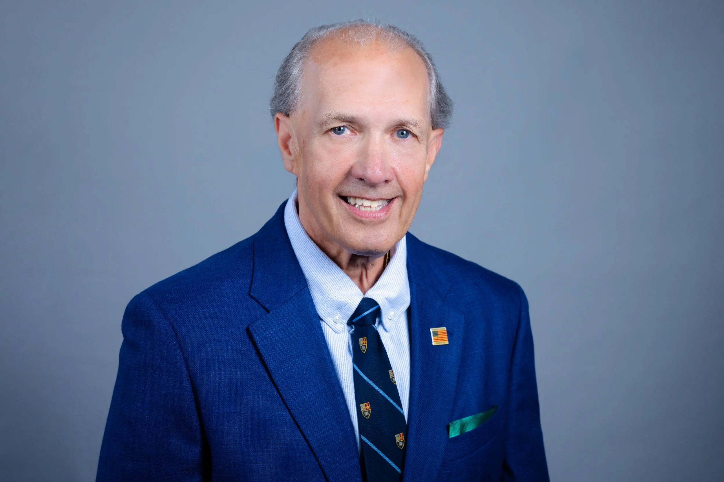 Portrait of an older man with gray hair wearing a blue suit, a light blue shirt, and a patterned tie, smiling at the camera.