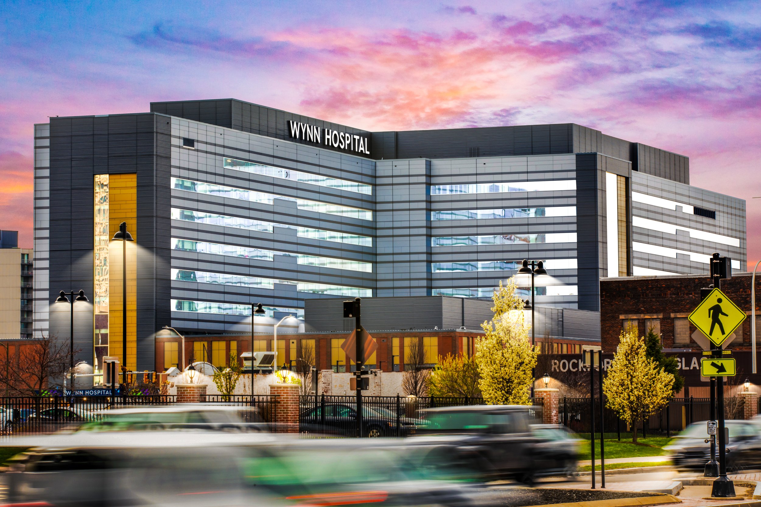 Modern hospital building with the sign 'Wynn Hospital' under a colorful sunset sky, busy street with moving cars and trees in foreground.