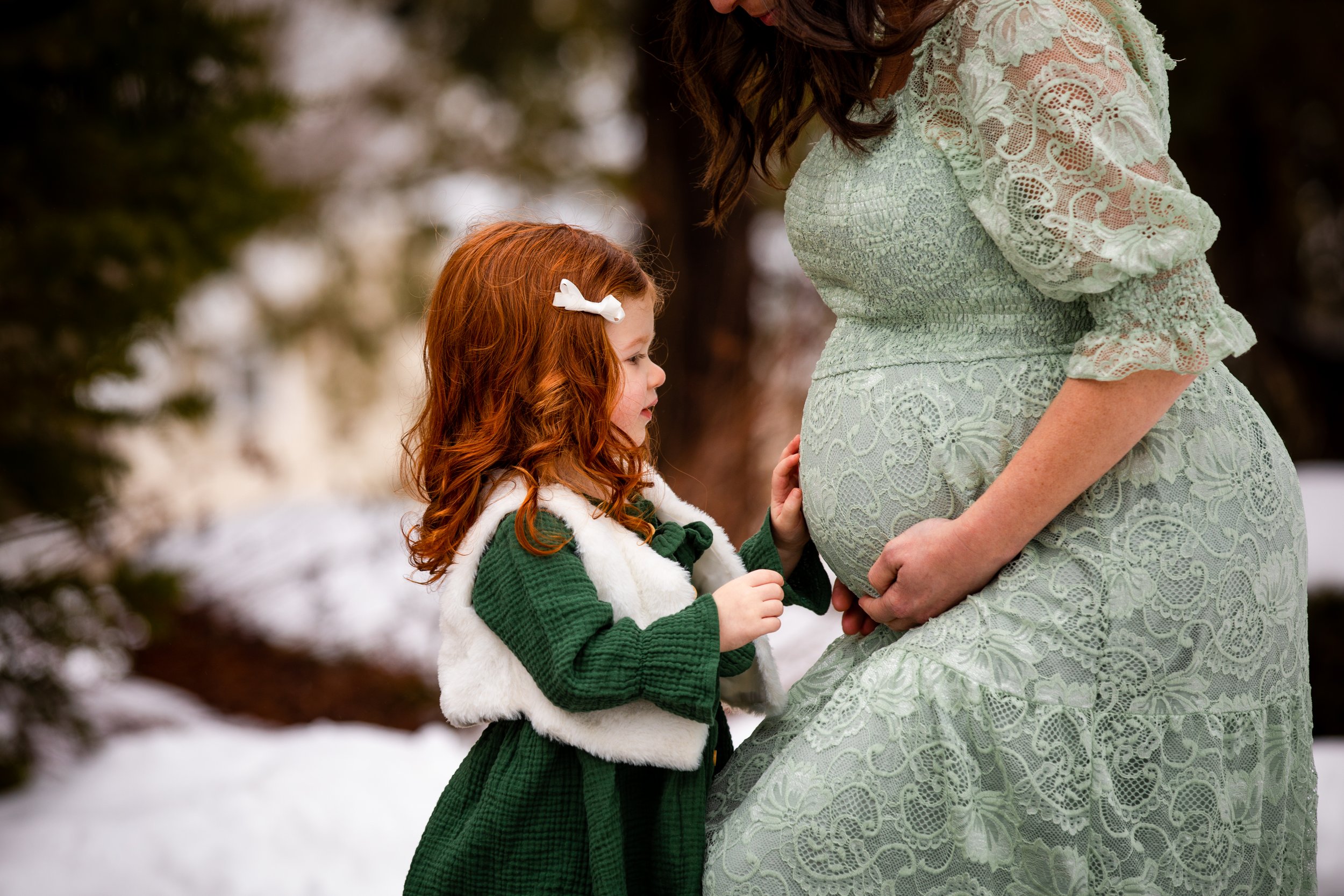 A young girl with red hair, wearing a green dress and a white fur vest, gently touching the pregnant belly of a woman in a light green lace dress outdoors in a snowy setting.
