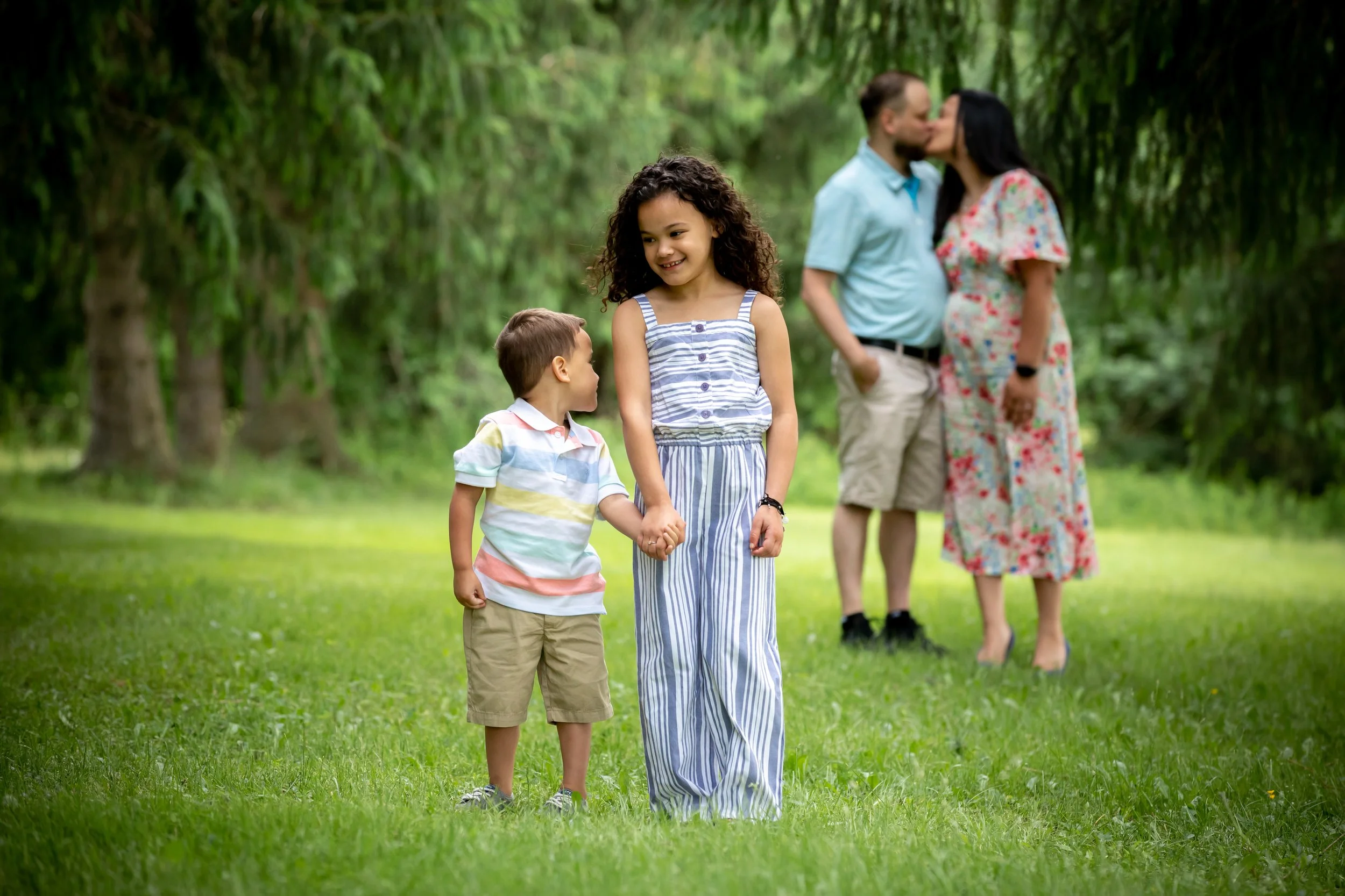 A family of four outdoors in a green park, with two children in the foreground holding hands and a man and woman in the background kissing.