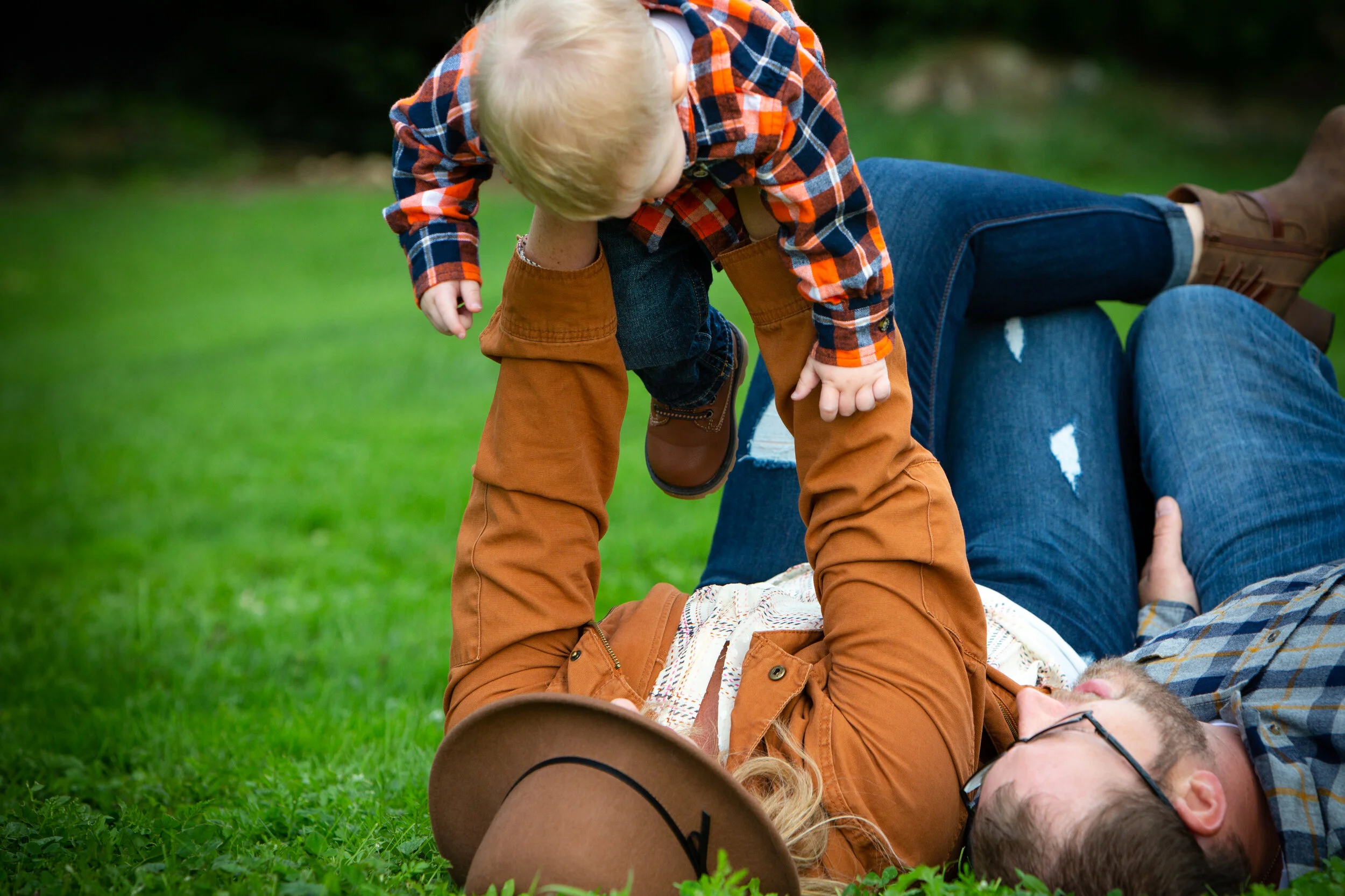 A child wearing a plaid shirt and brown pants is playing on an adult lying on the grass, seemingly wrestling or playing a game outdoors.