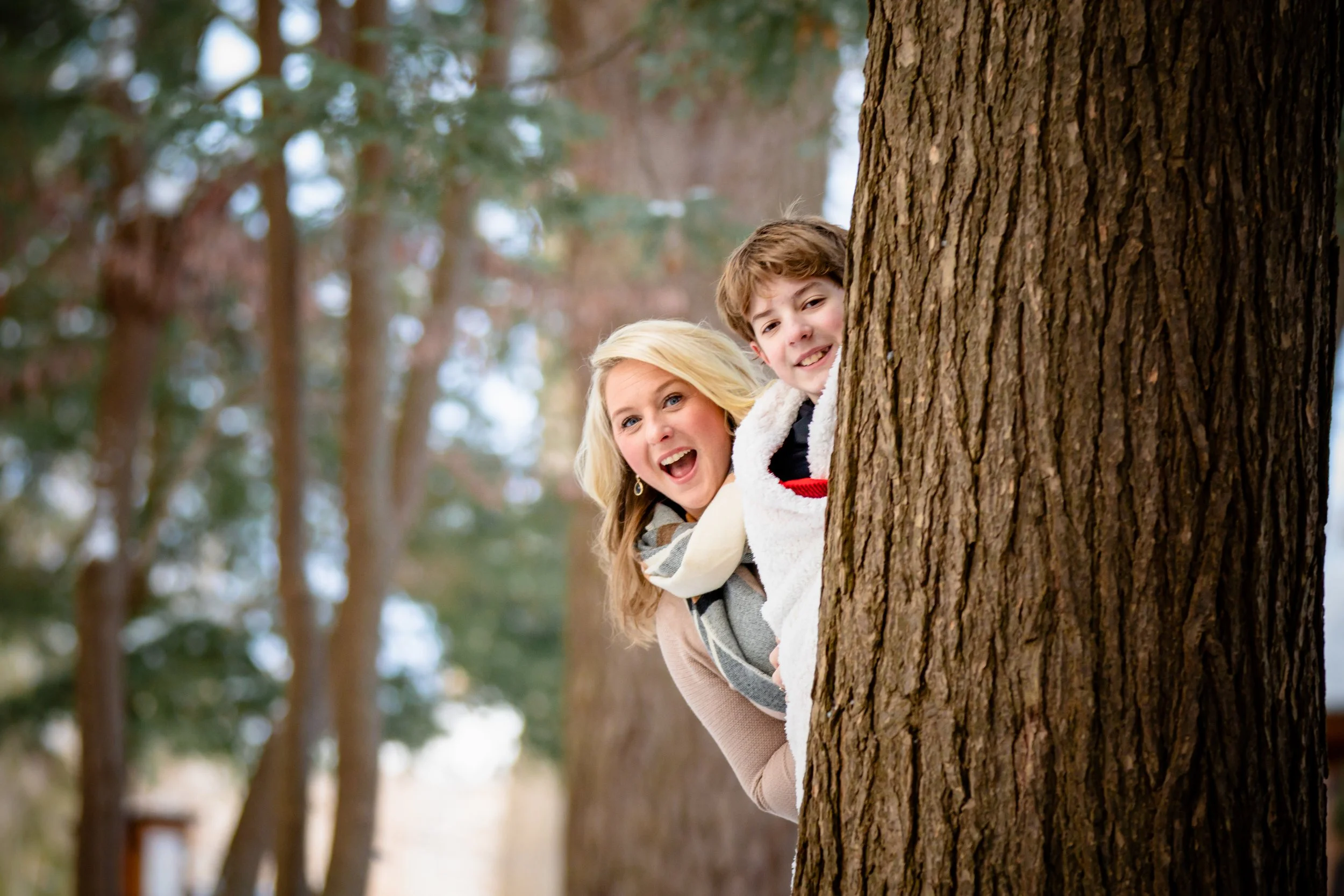 A woman and a boy peeking out from behind a large tree trunk in a wooded area, smiling and having fun.