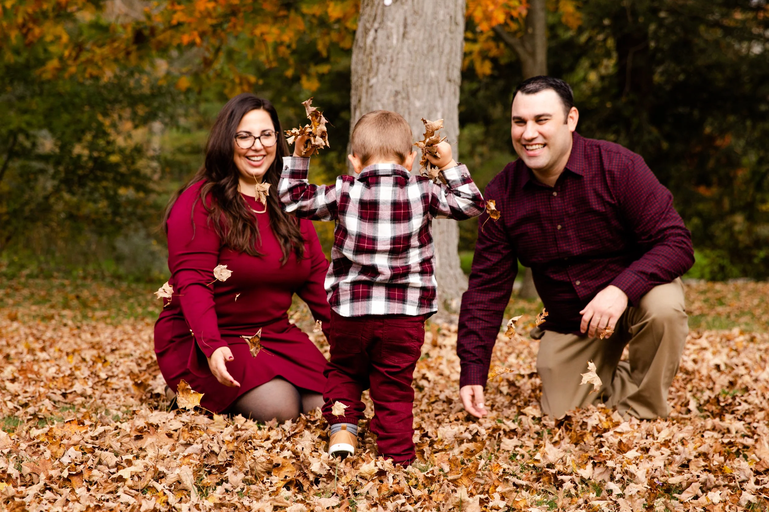 A family of three playing in a park covered with fallen autumn leaves. The woman and man are kneeling while the child throws leaves into the air. The background includes trees with autumn foliage.