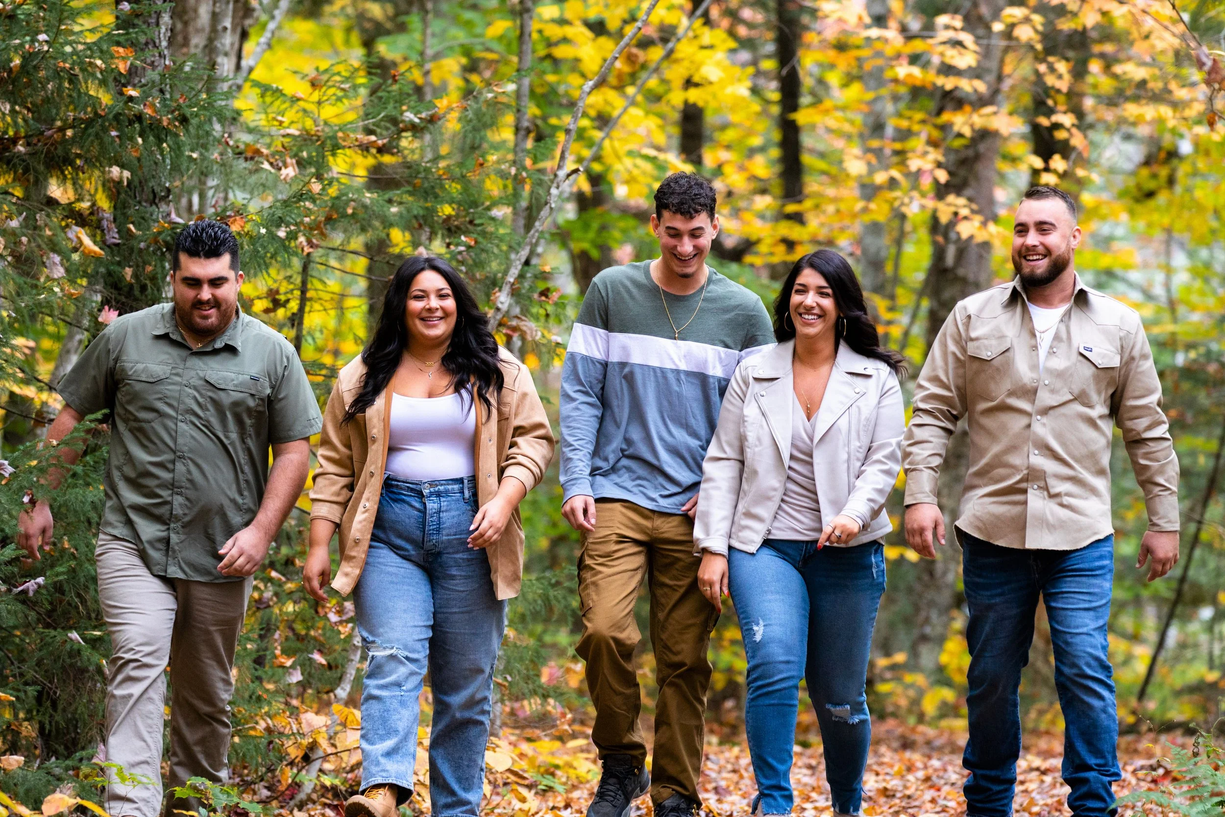 A group of five friends walking through a forest with fall foliage, smiling and enjoying each other's company.