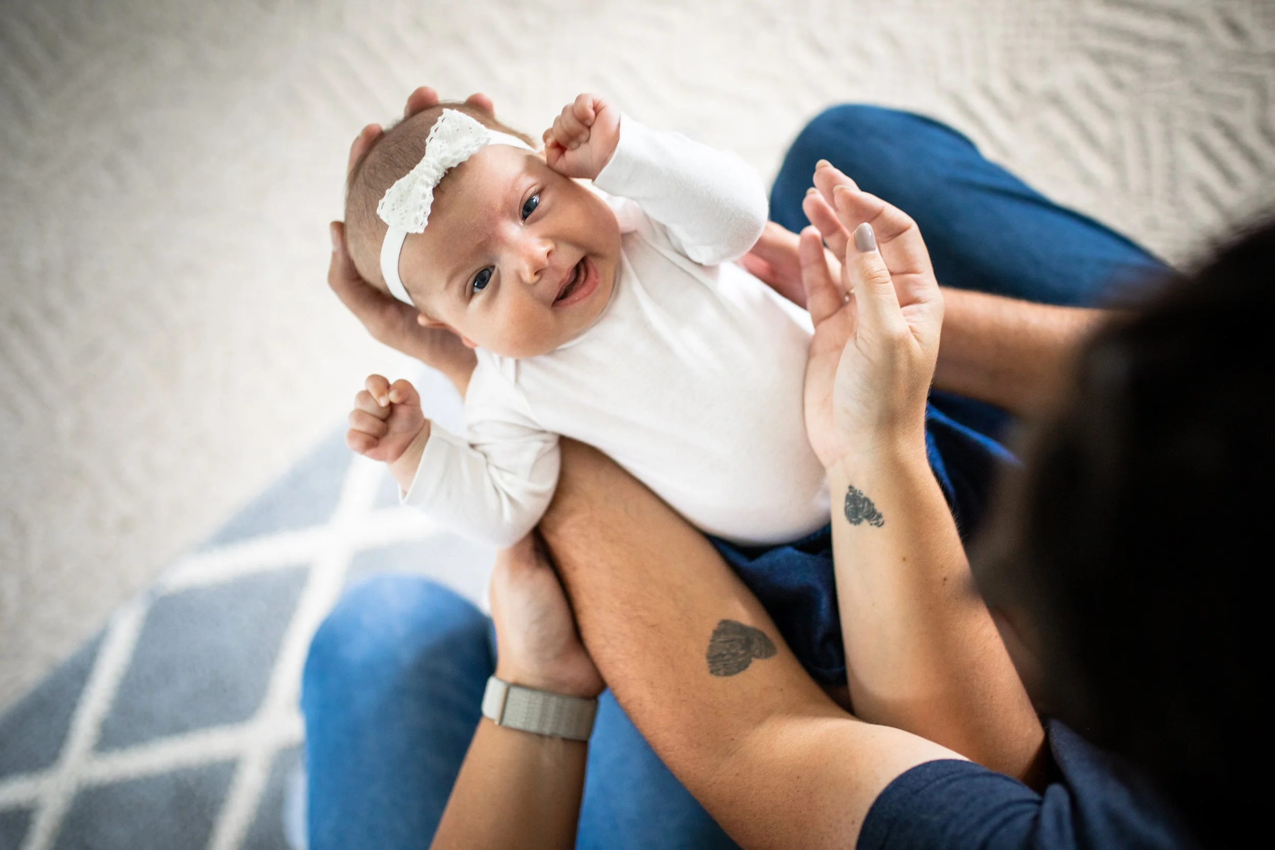 A smiling baby with a headband lying on an adult's lap, being gently held by the adult's hands. The adult has tattoos on their forearms and is wearing a watch. The background shows a patterned rug.