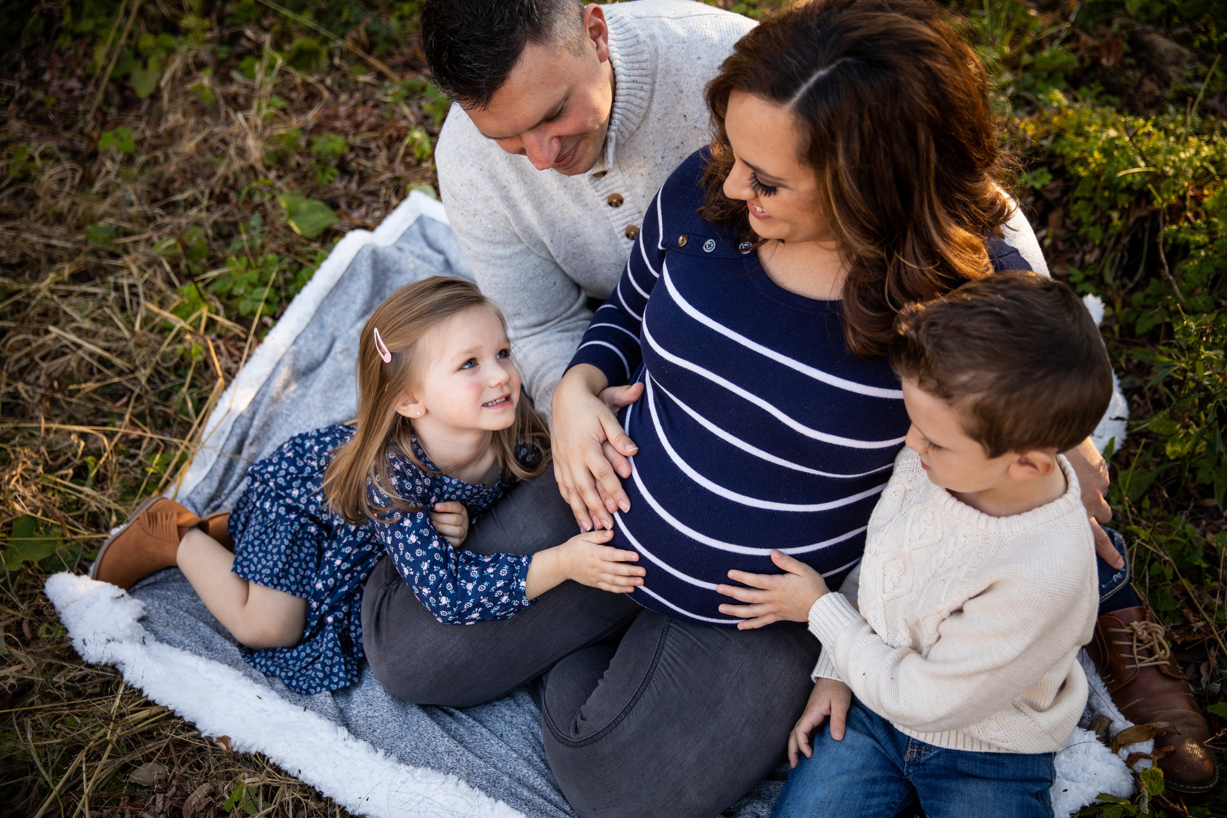 A pregnant woman sits on a blanket outdoors with her family, including a young girl and a young boy, who are touching her baby bump and looking at her affectionately.