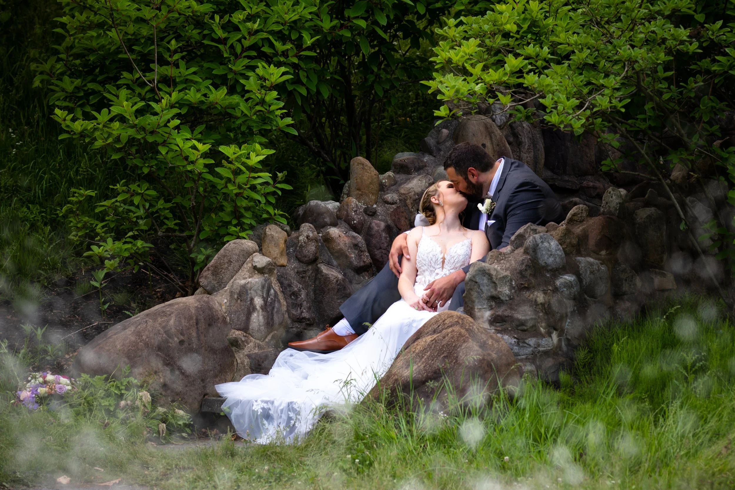A bride and groom sit among rocks and greenery, sharing a kiss.