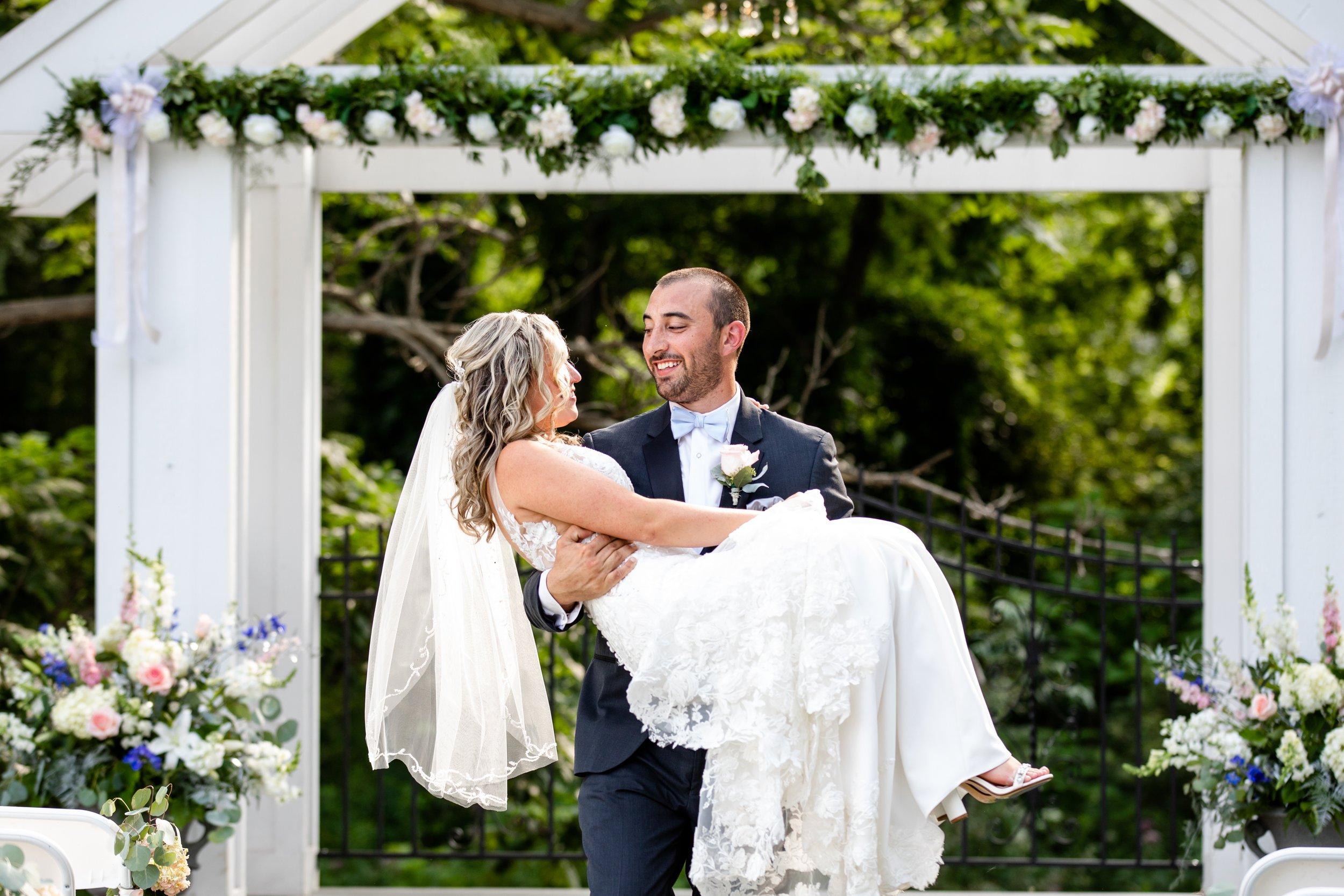 A groom carrying a bride in a white wedding dress under a floral wedding arch outdoors with green trees in the background.
