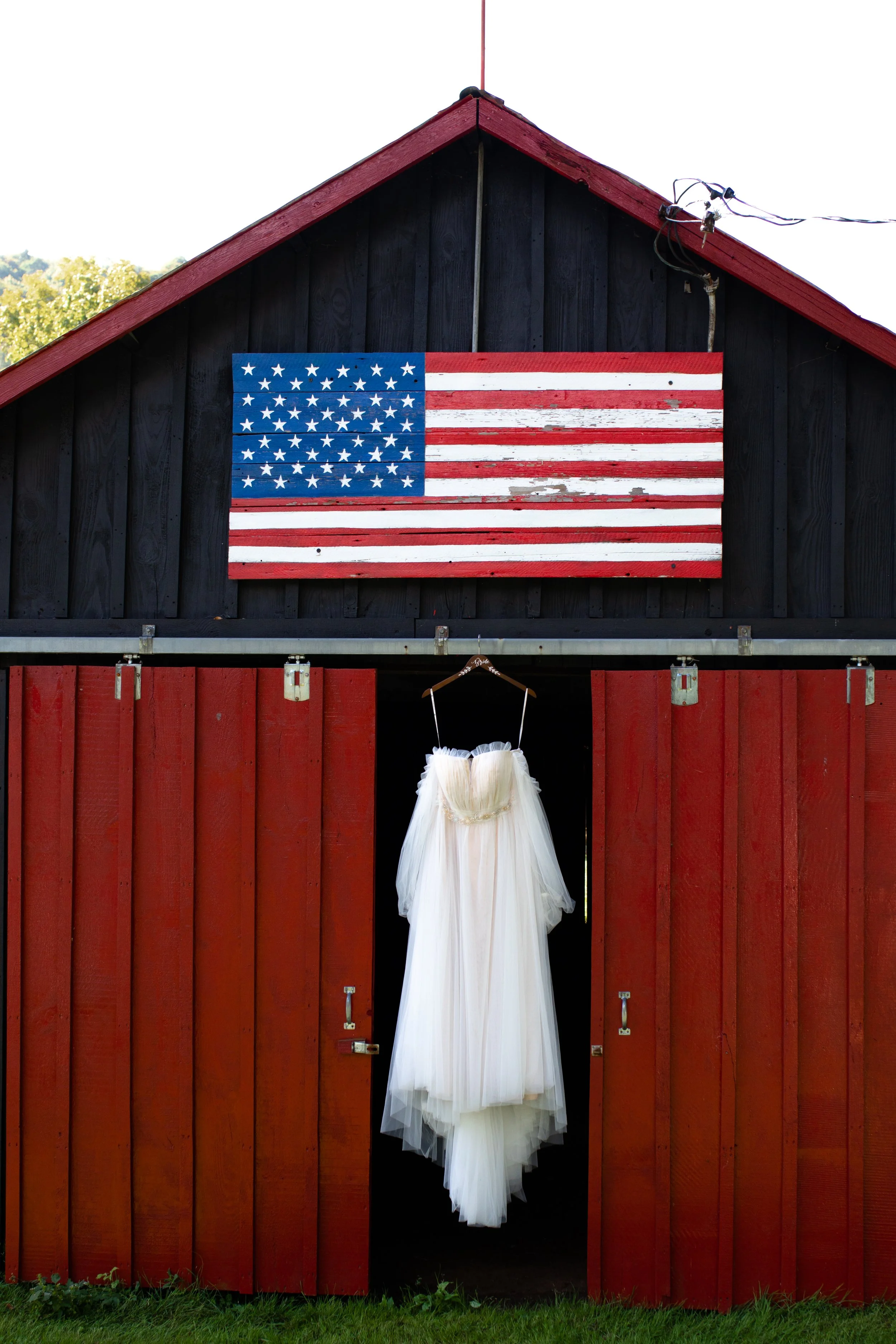 A white wedding dress hanging inside a red barn with black walls, with an American flag mounted on the exterior wall above.