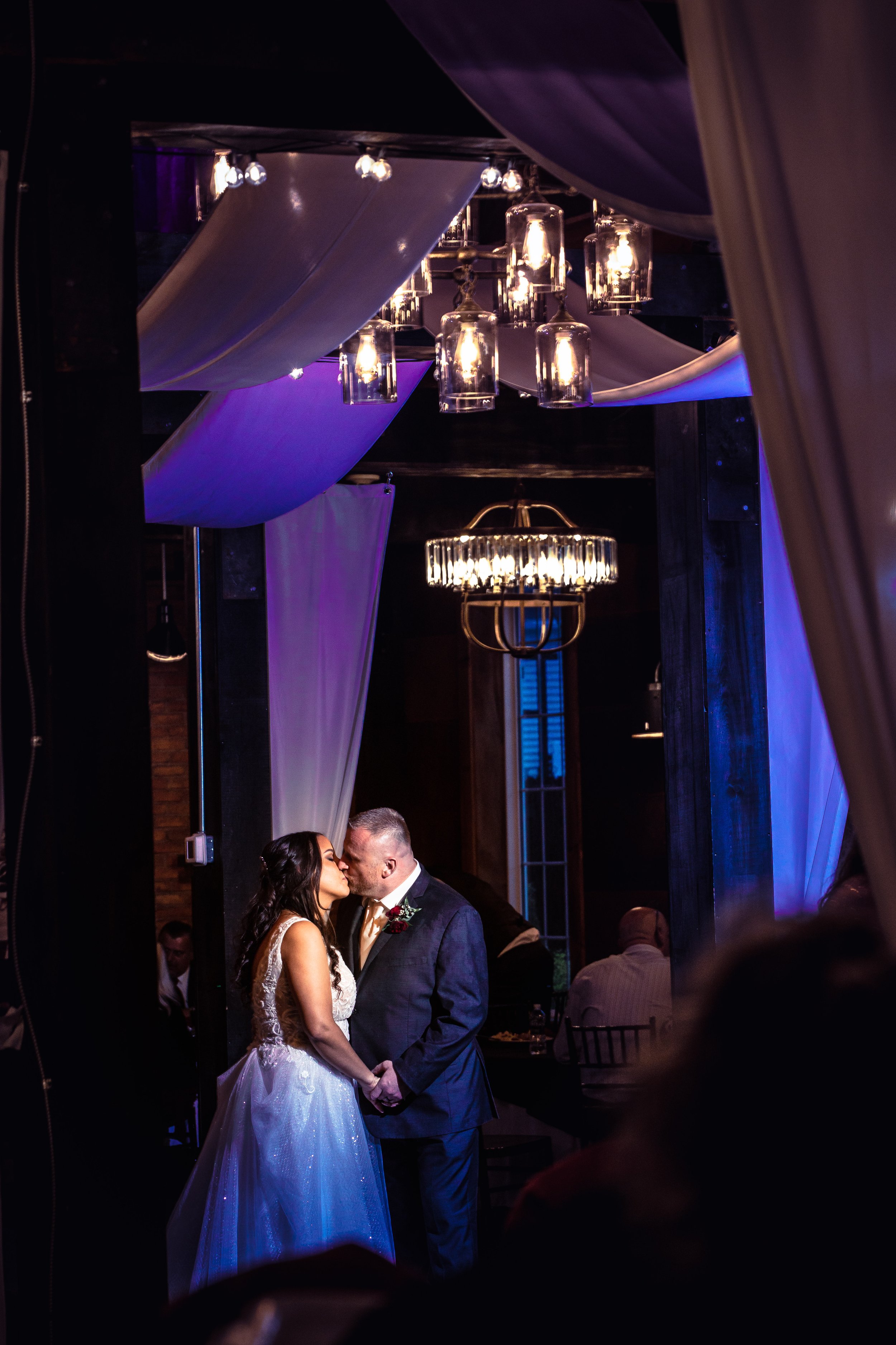 A bride and groom sharing a kiss during their wedding ceremony, with dim lighting, hanging glass light fixtures, and draped white fabric decorating the ceiling, creating an intimate atmosphere.
