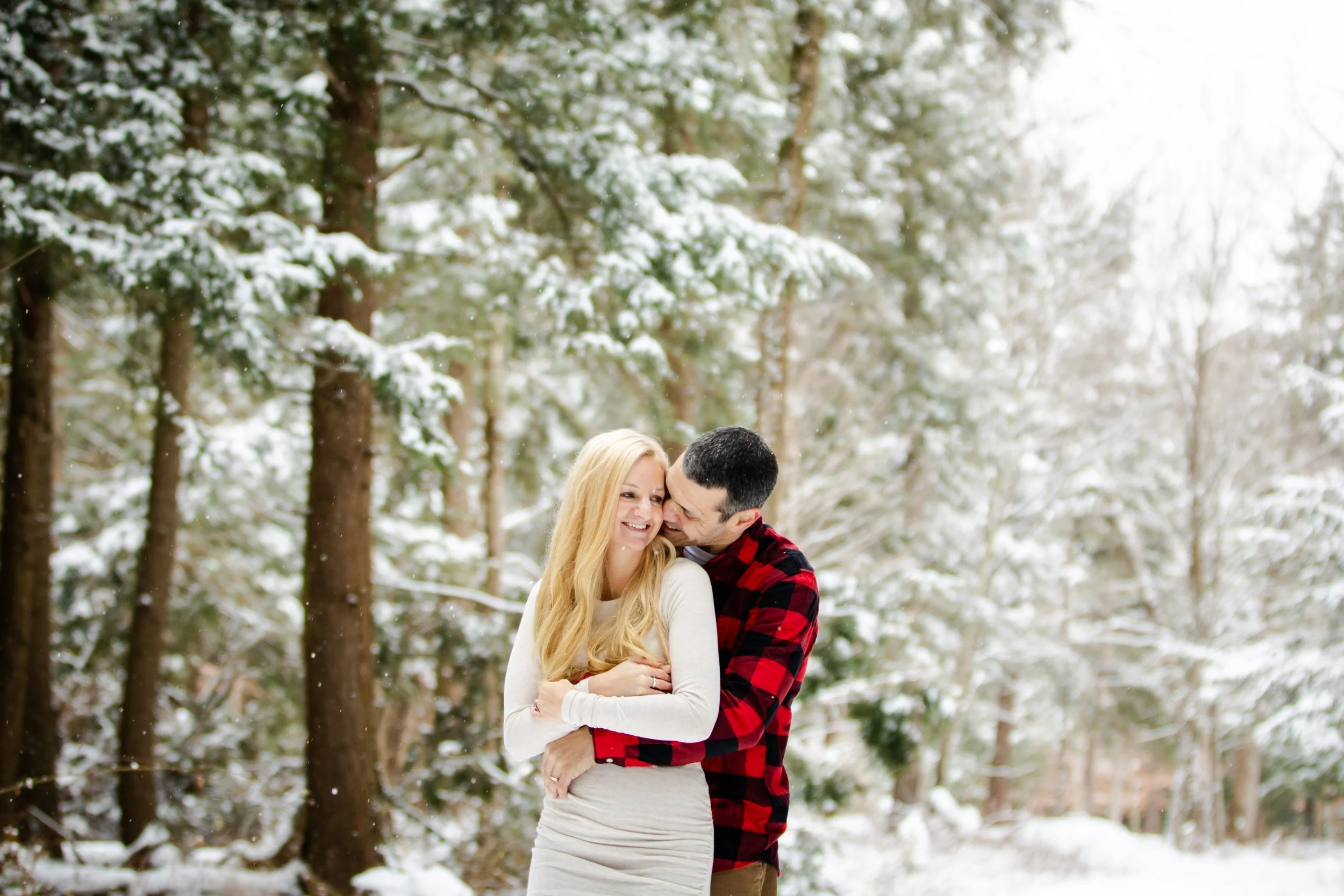 A couple embracing in a snowy forest, with snow-covered trees in the background.