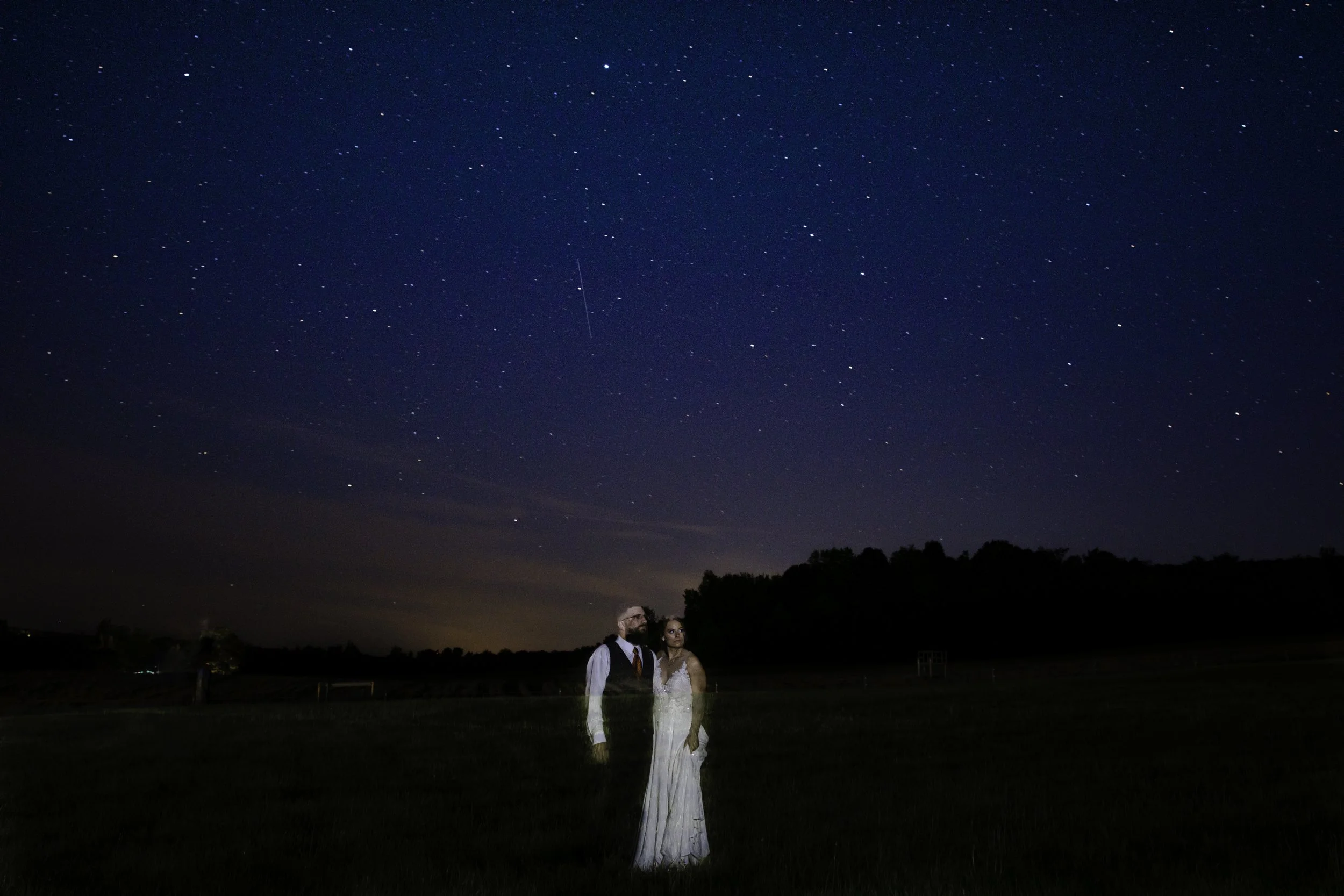 A couple standing on grass at night under a star-filled sky, with faint clouds and a shooting star visible.