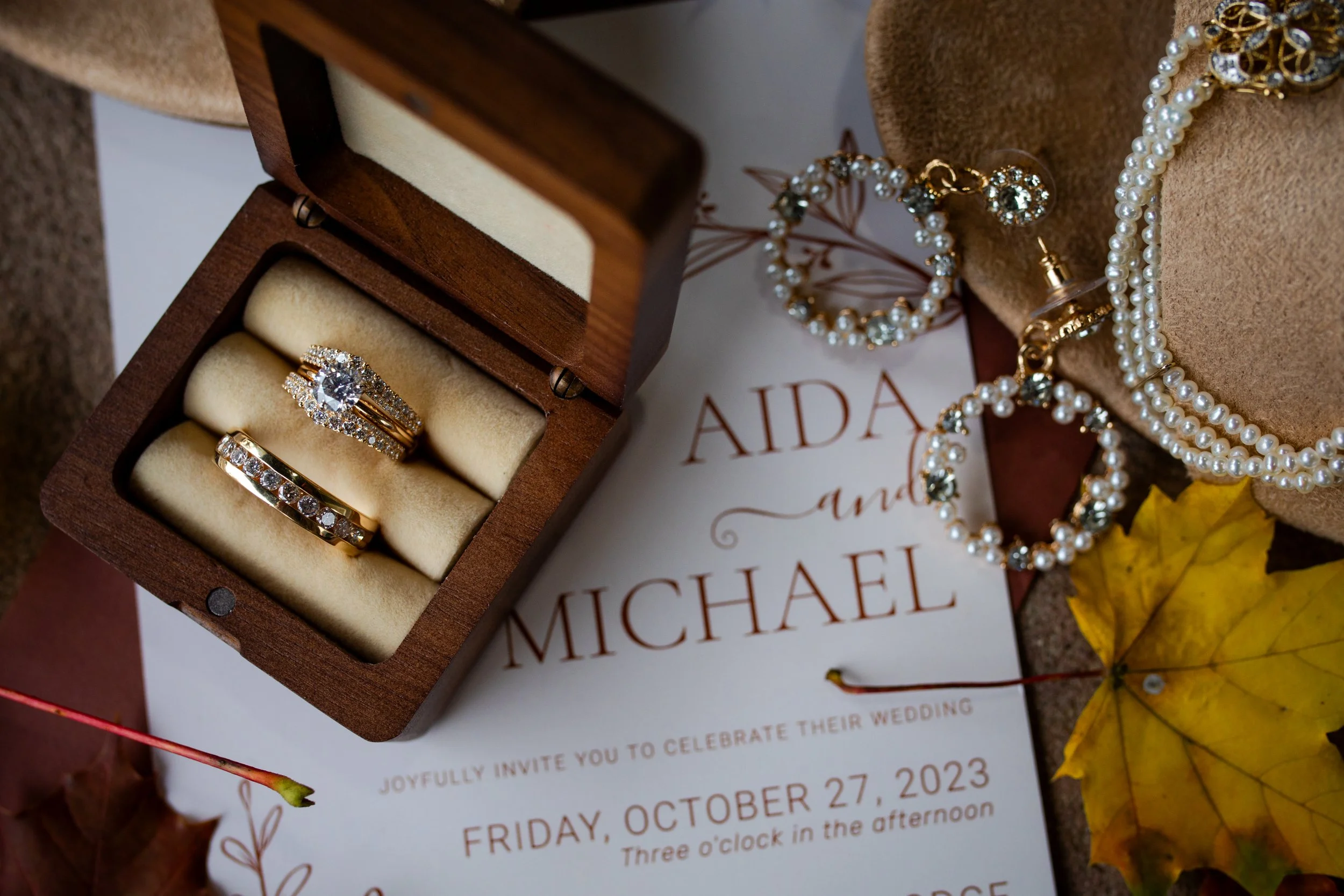 Wedding rings in a wooden box surrounded by jewelry, a wedding invitation, and fall leaves.