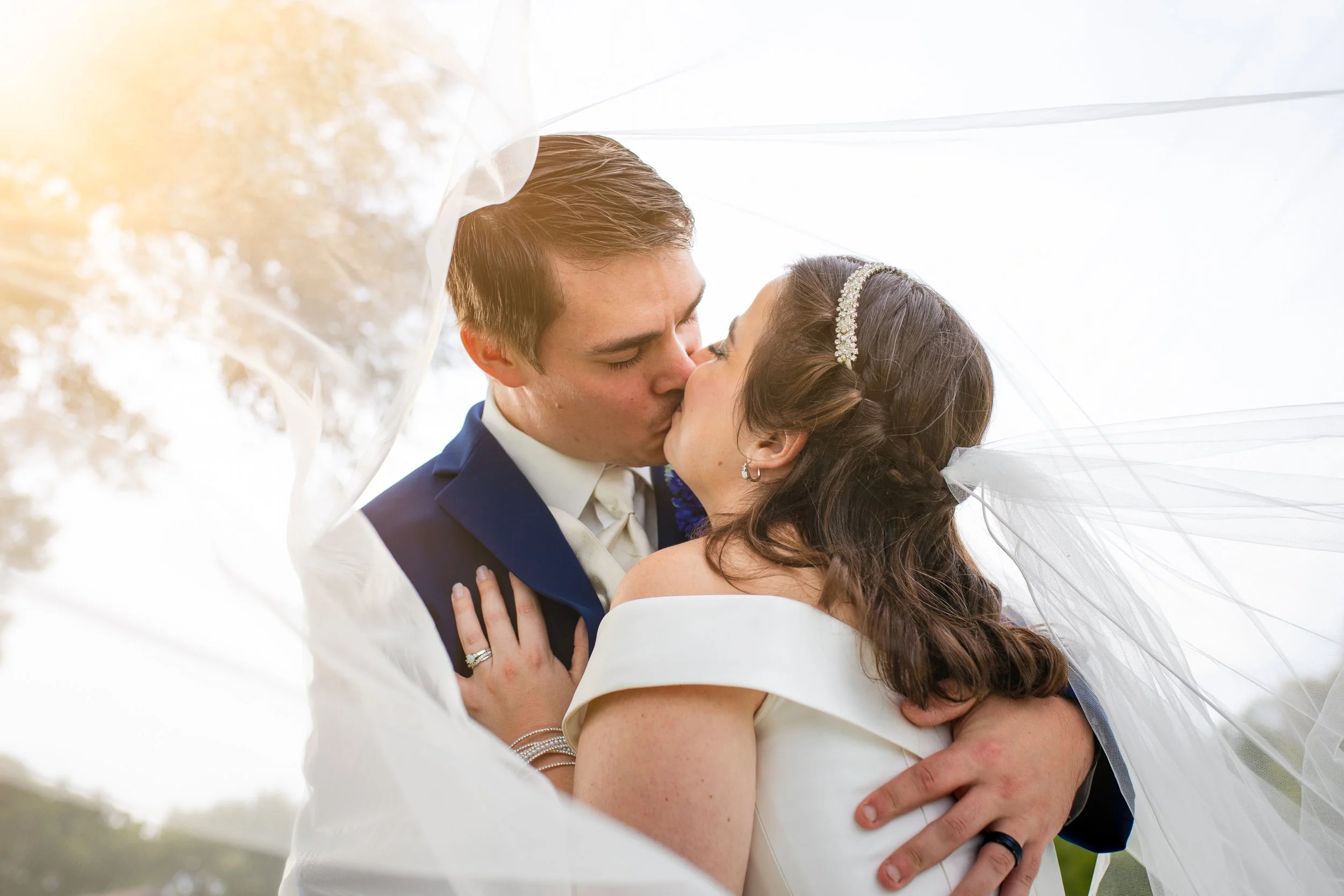 Bride and groom kissing under wedding veil outdoors, wedding attire, sunlight, romantic moment.