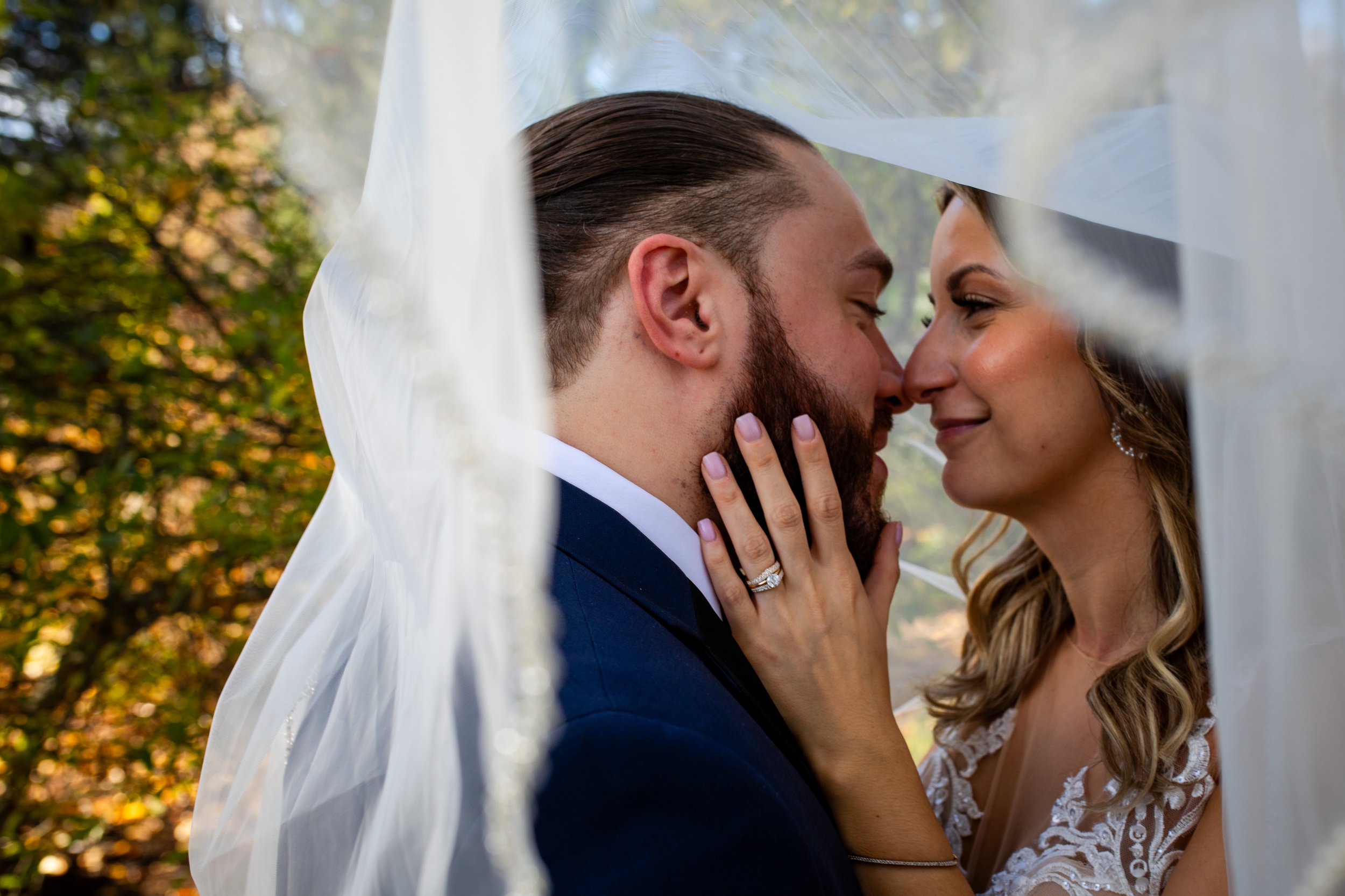 A bride and groom touching noses and smiling, with the bride's hand on the groom's beard, behind a sheer white veil, outdoor setting with trees in the background.