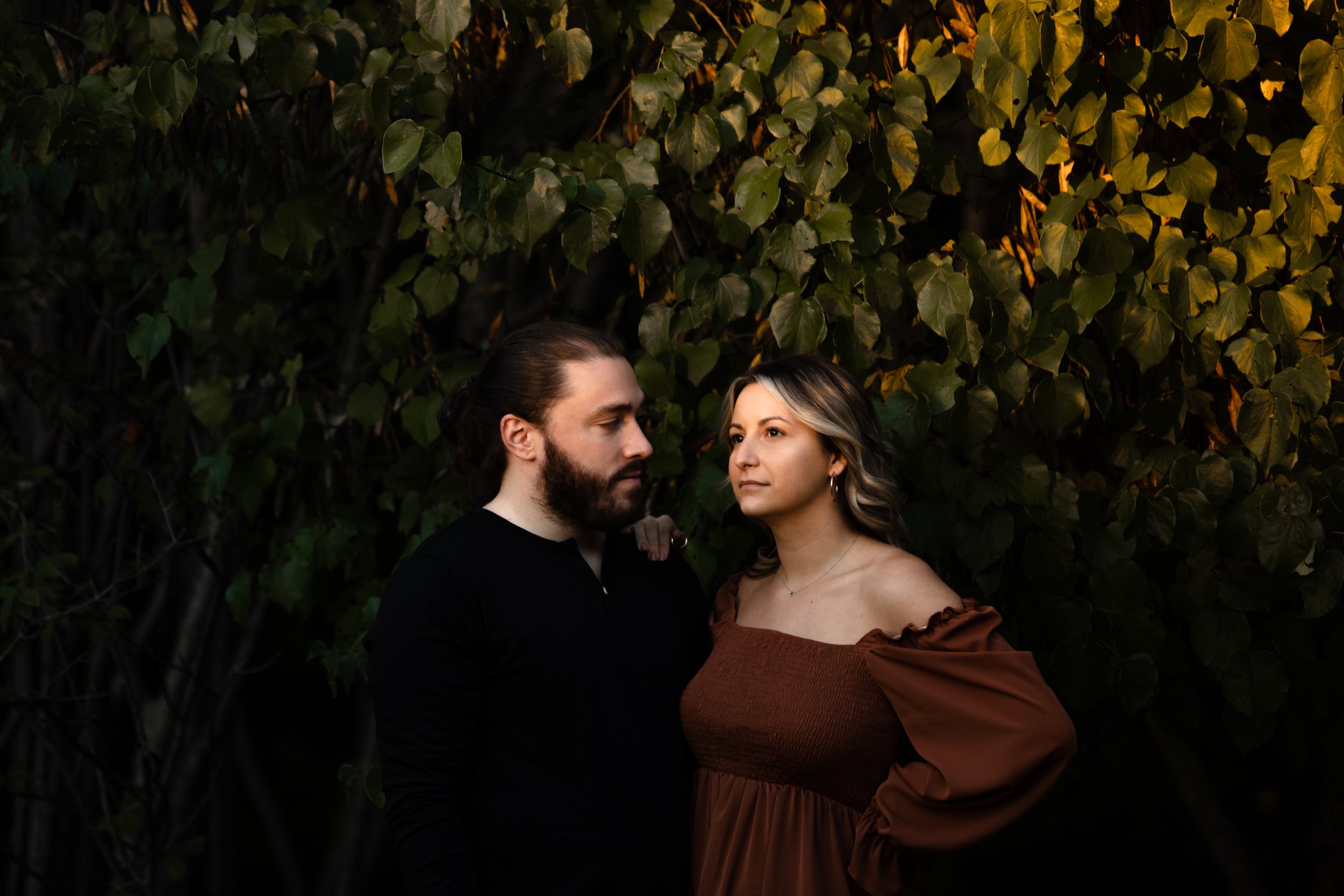 A man with long hair and a beard wearing a black shirt and a woman with shoulder-length hair wearing a brown off-shoulder dress, standing close to each other outdoors against a backdrop of green leafy plants.