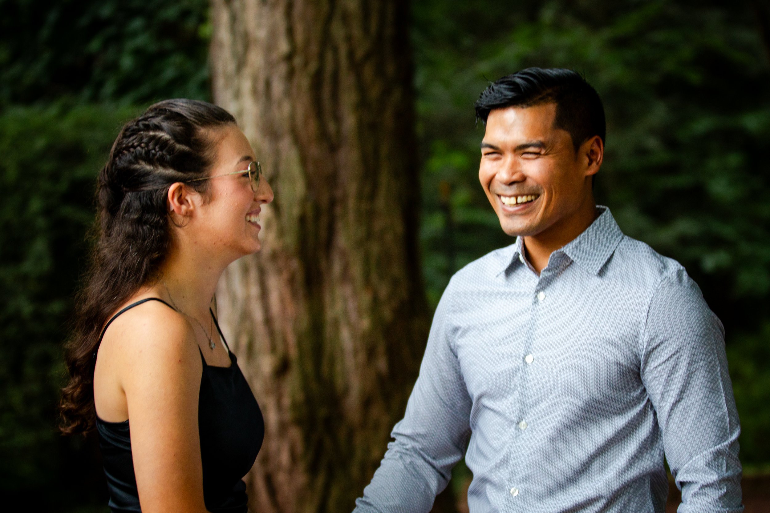 A smiling couple talking outdoors in front of a large tree with green foliage in the background.