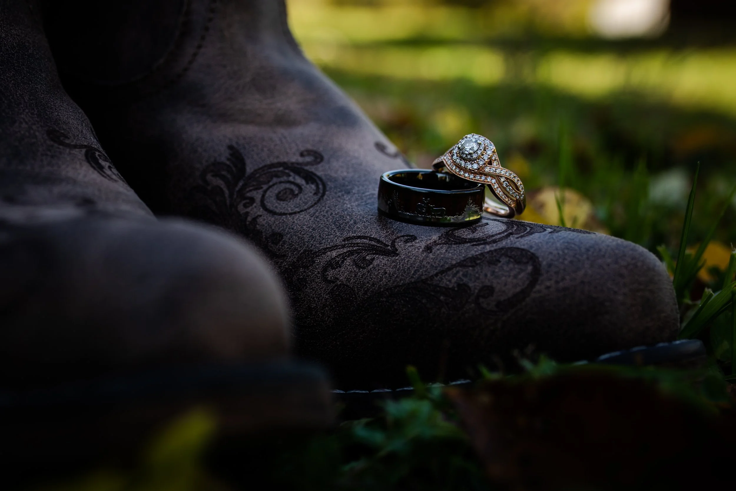 Engagement rings resting on a person's dark floral-patterned clothing outdoors with grass and fallen leaves in the background.