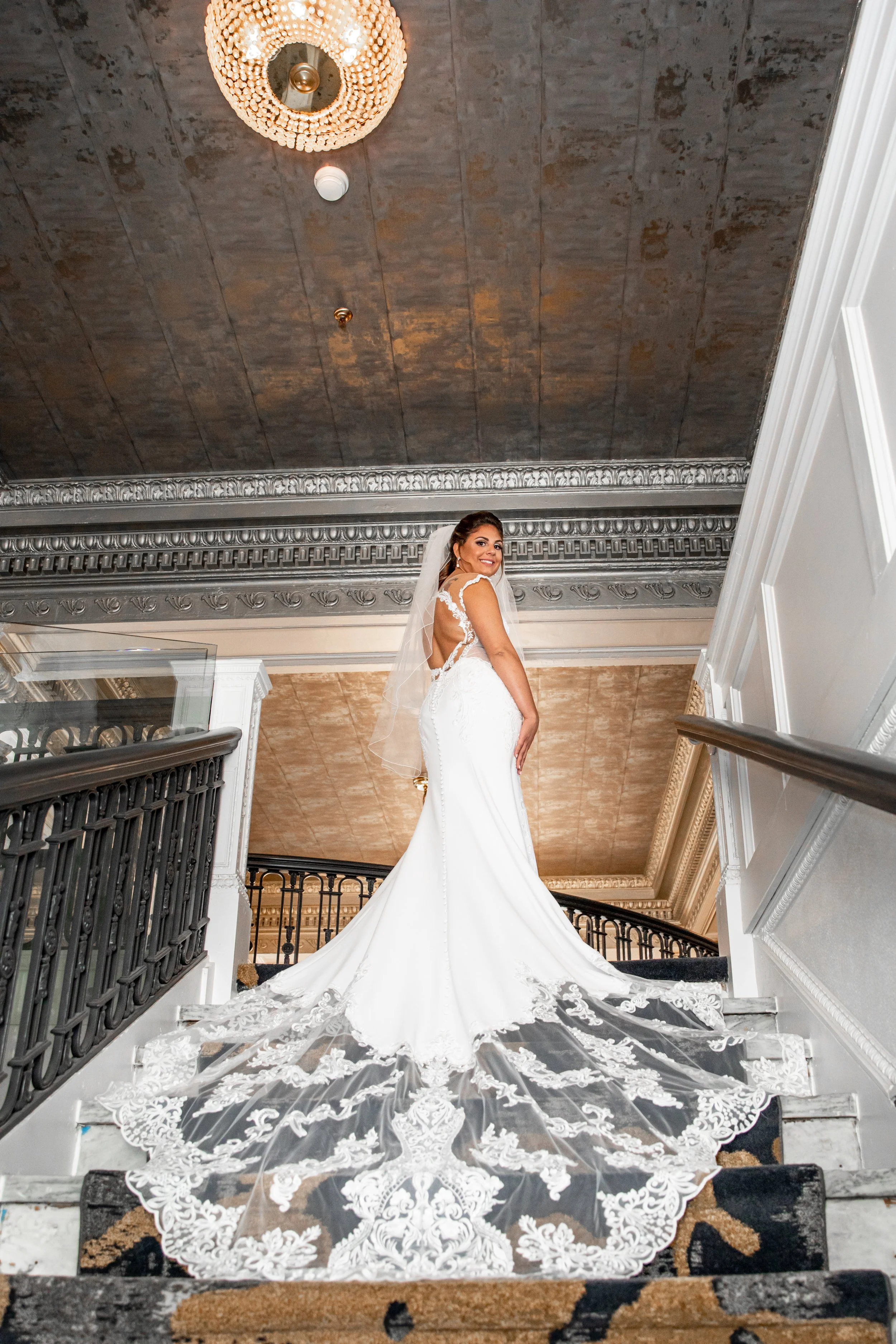 Bride in a white wedding dress with lace details, standing on a staircase, smiling at the camera with a veil, ornate ceiling and railing in the background.