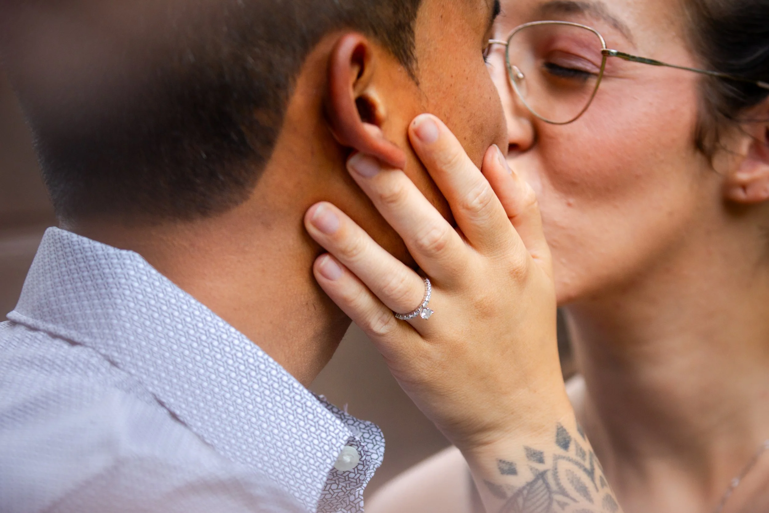Close-up of a couple kissing, woman with glasses and a diamond ring, hugging a man with short hair, wearing a gray collared shirt.