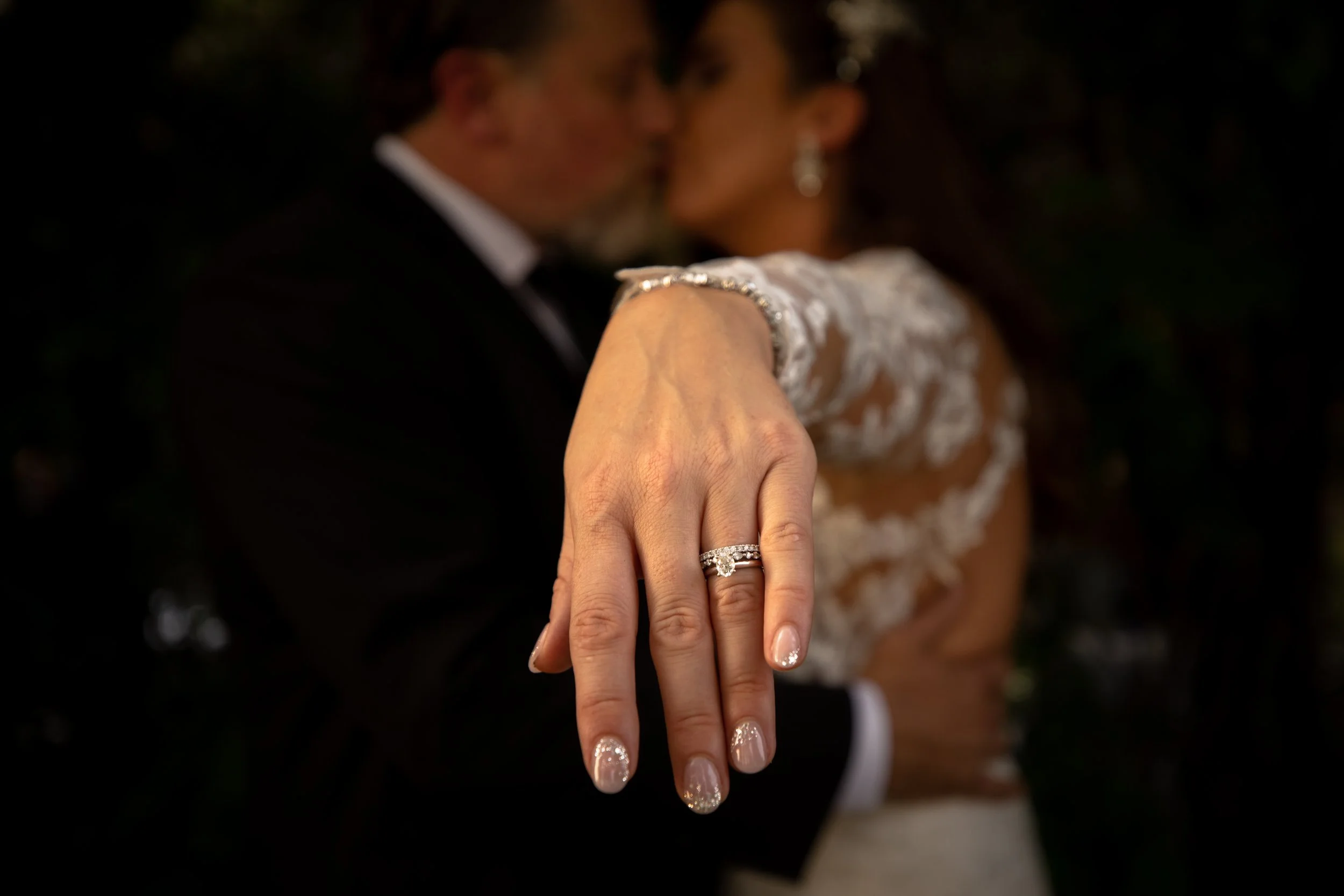 Close-up of a woman's hand showing a wedding or engagement ring, with the couple kissing in the background.