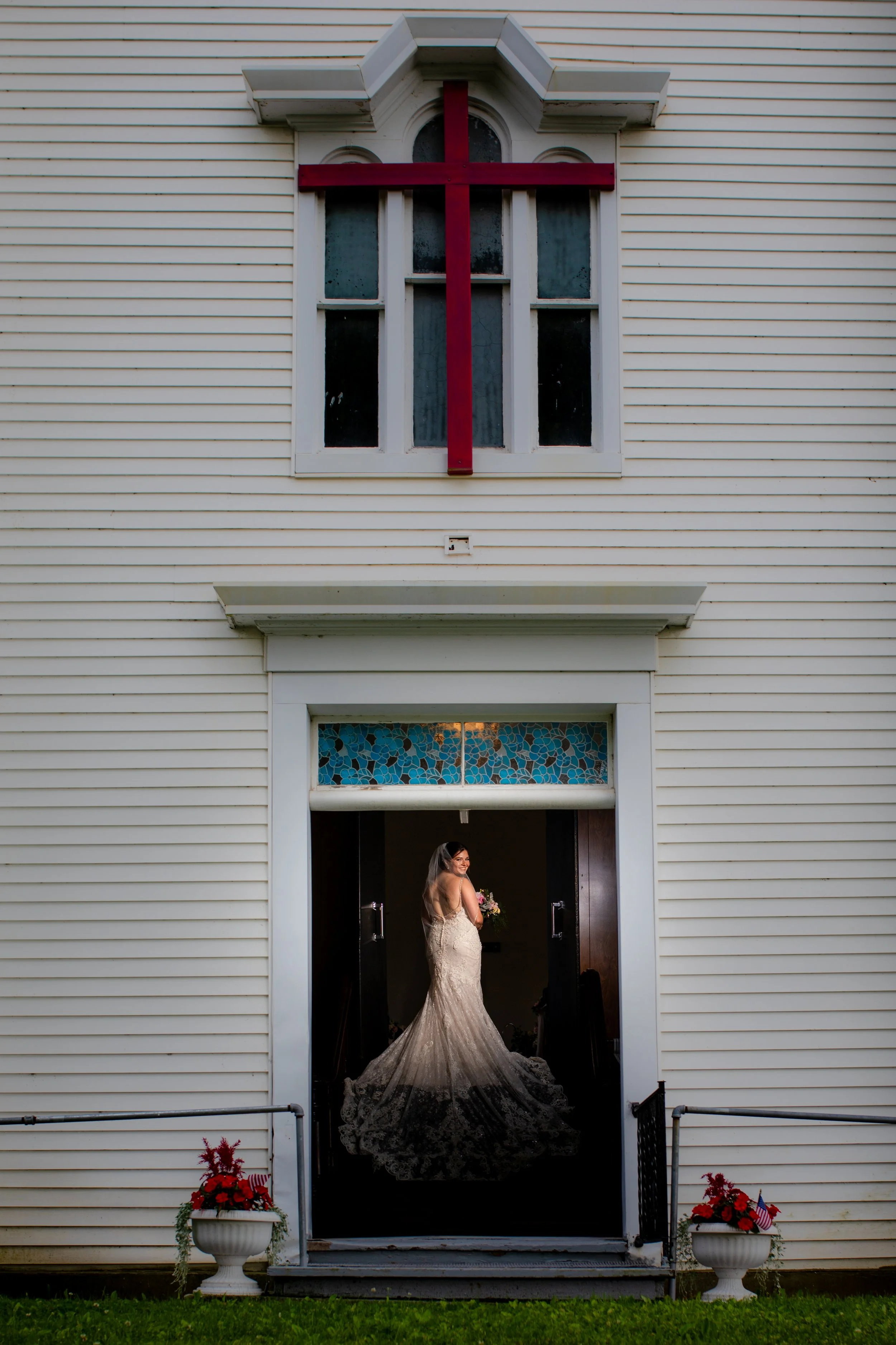 A bride in a wedding dress standing inside a church doorway, looking back and smiling, with the church painted white and decorated with patriotic flowers outside.