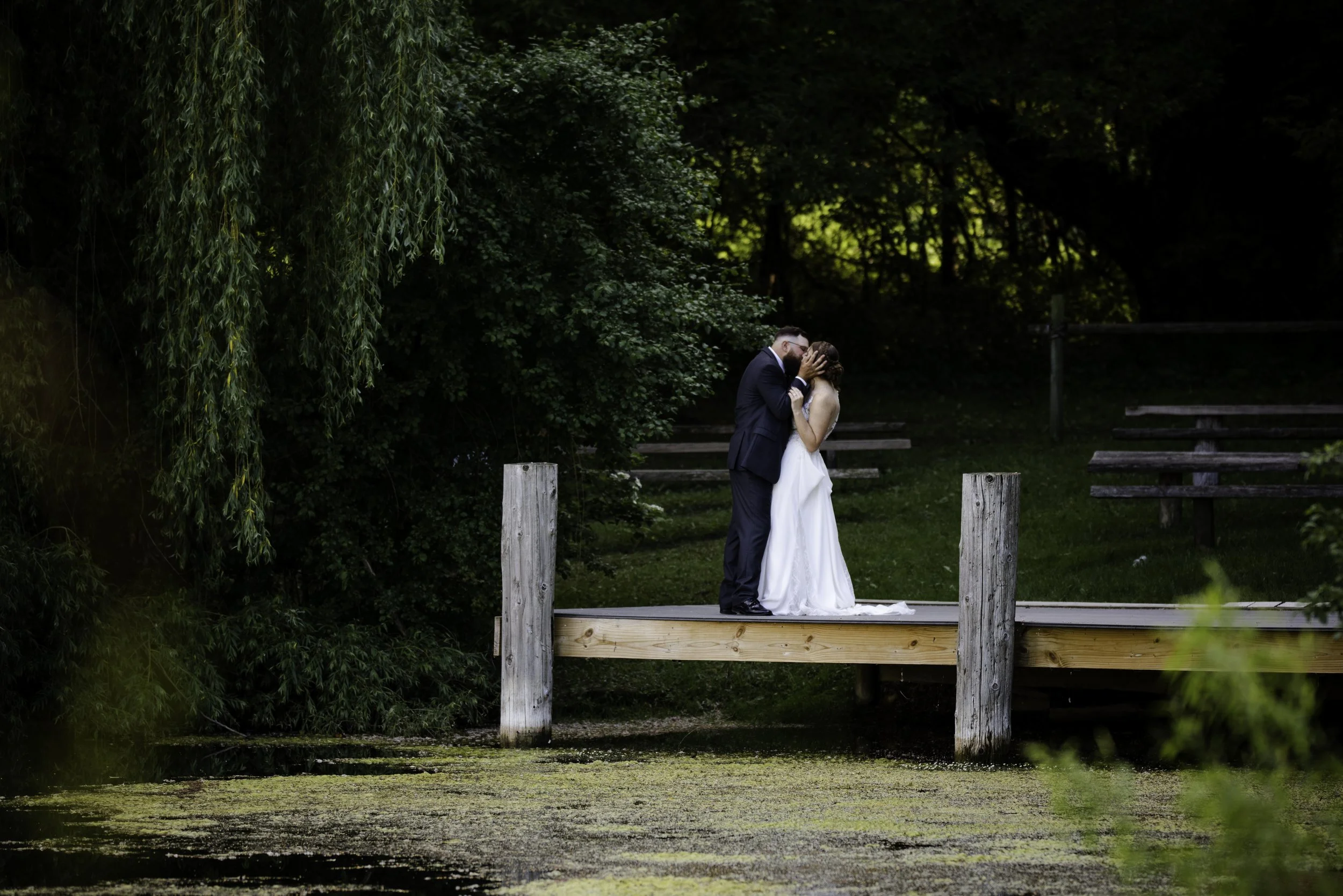 A bride and groom sharing a kiss on a wooden dock by a pond surrounded by trees.
