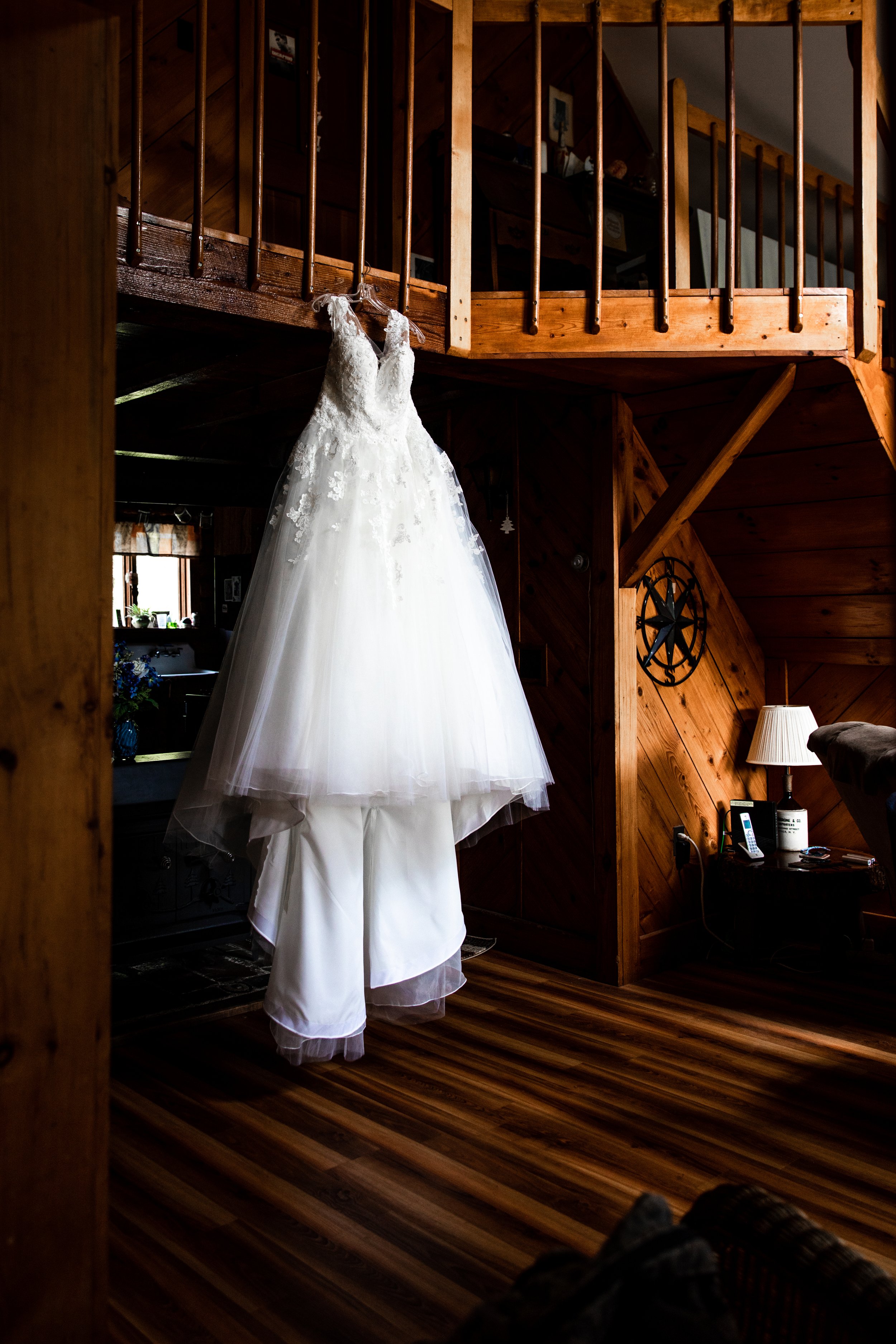 A wedding dress hanging from the wooden railing of a loft in a cozy, wood-paneled room. The dress is white with lace details, and the room features wooden furniture and warm lighting.