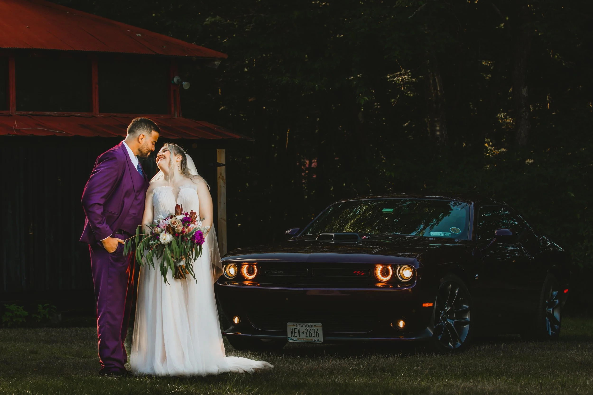 A bride and groom stand close together, smiling and looking at each other, in front of a black muscle car with glowing headlights, outdoors during dusk or evening.