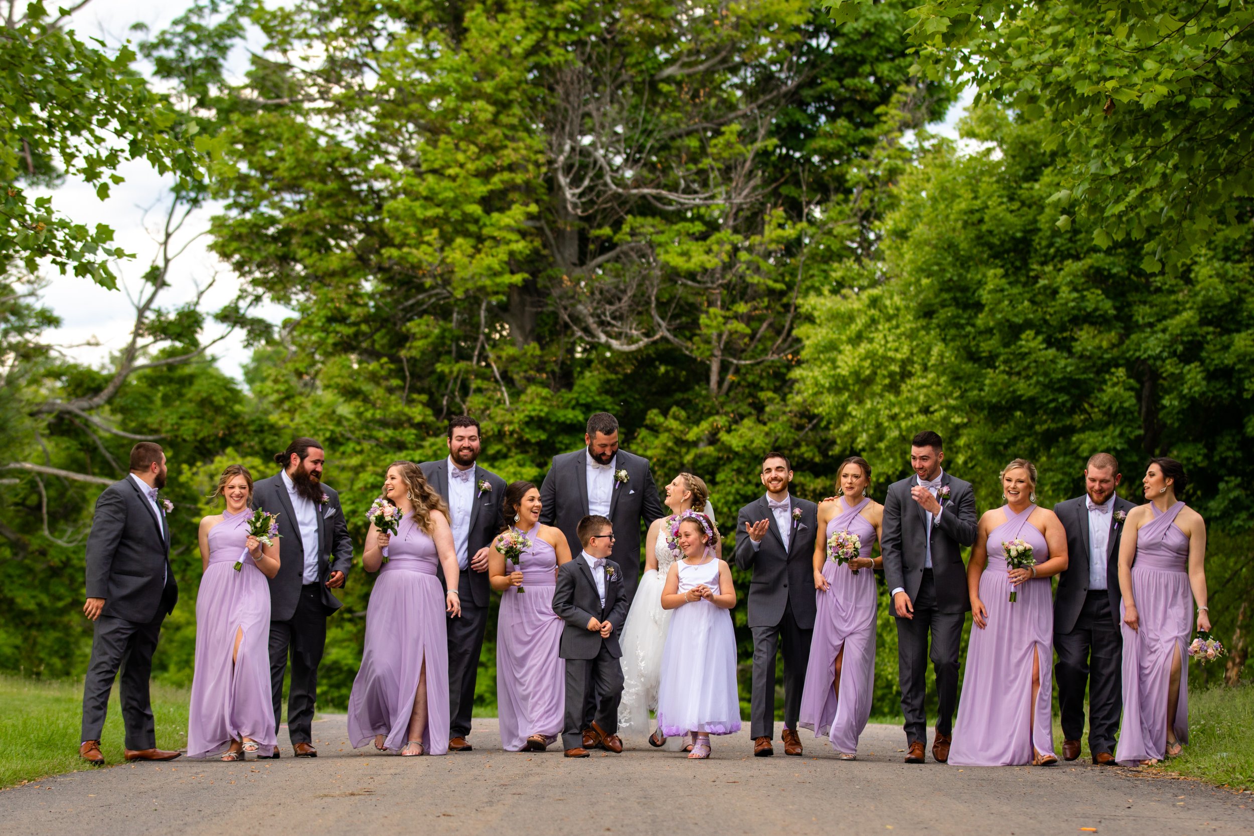 Group of wedding guests, including bridesmaids in lavender dresses and groomsmen in suits, walking outdoors on a paved path surrounded by lush green trees.