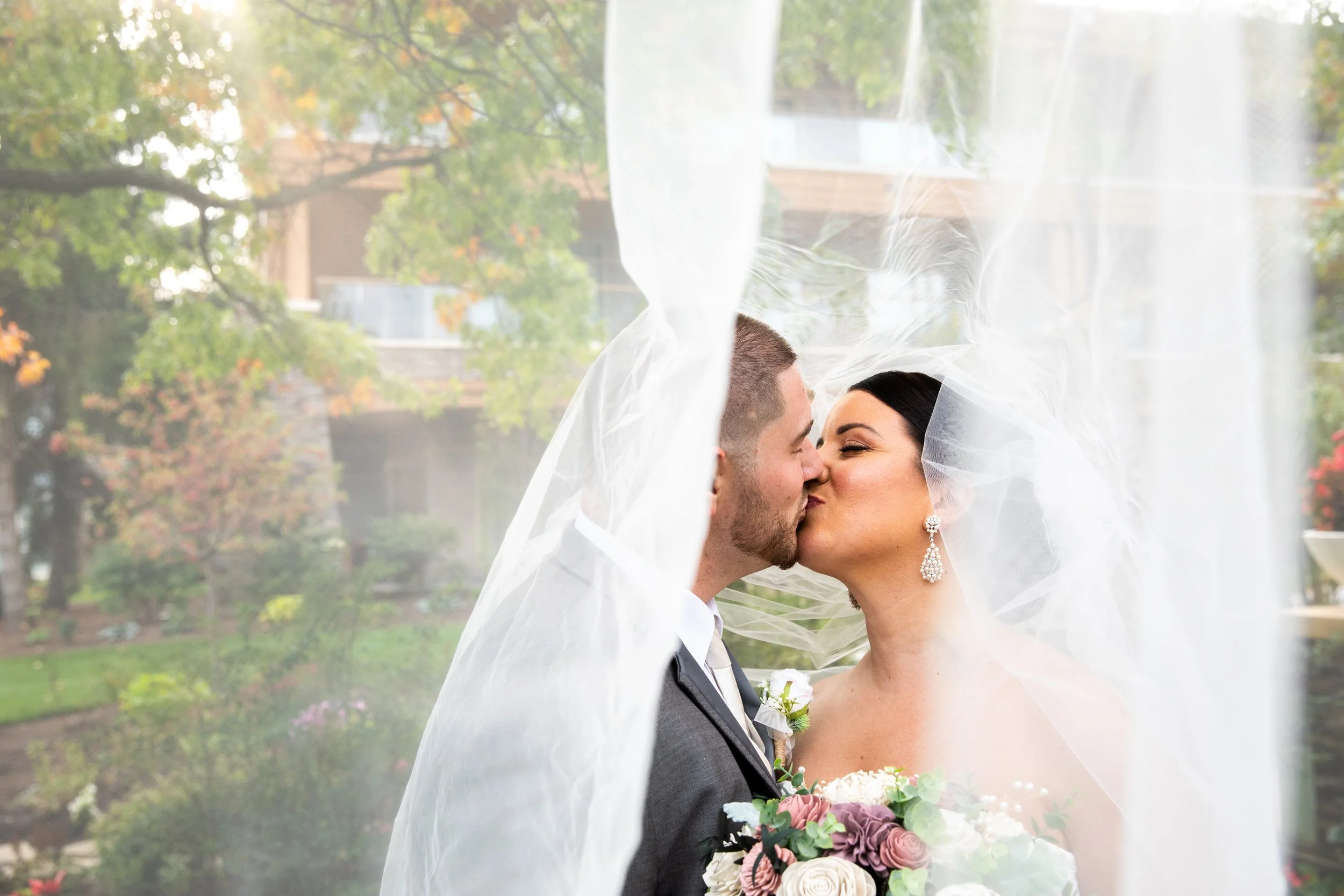 Bride and groom sharing a kiss behind a wedding veil, outdoors with trees and buildings in the background.