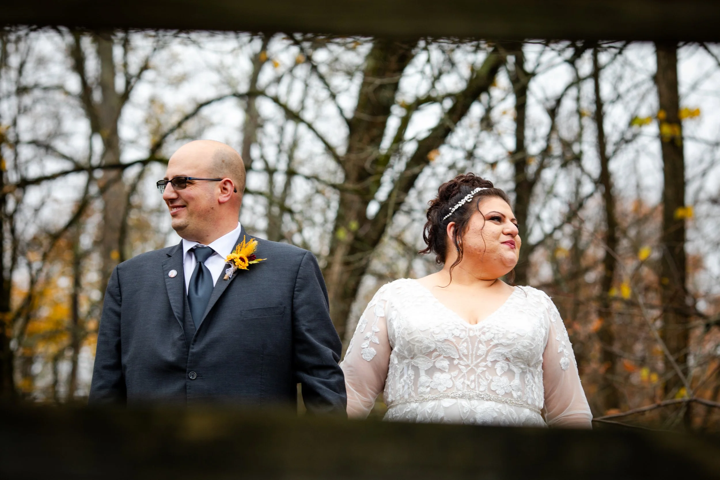 A couple on their wedding day outdoors, the groom in a dark suit with a sunflower boutonniere and the bride in a white lace gown, standing in front of leafless trees.