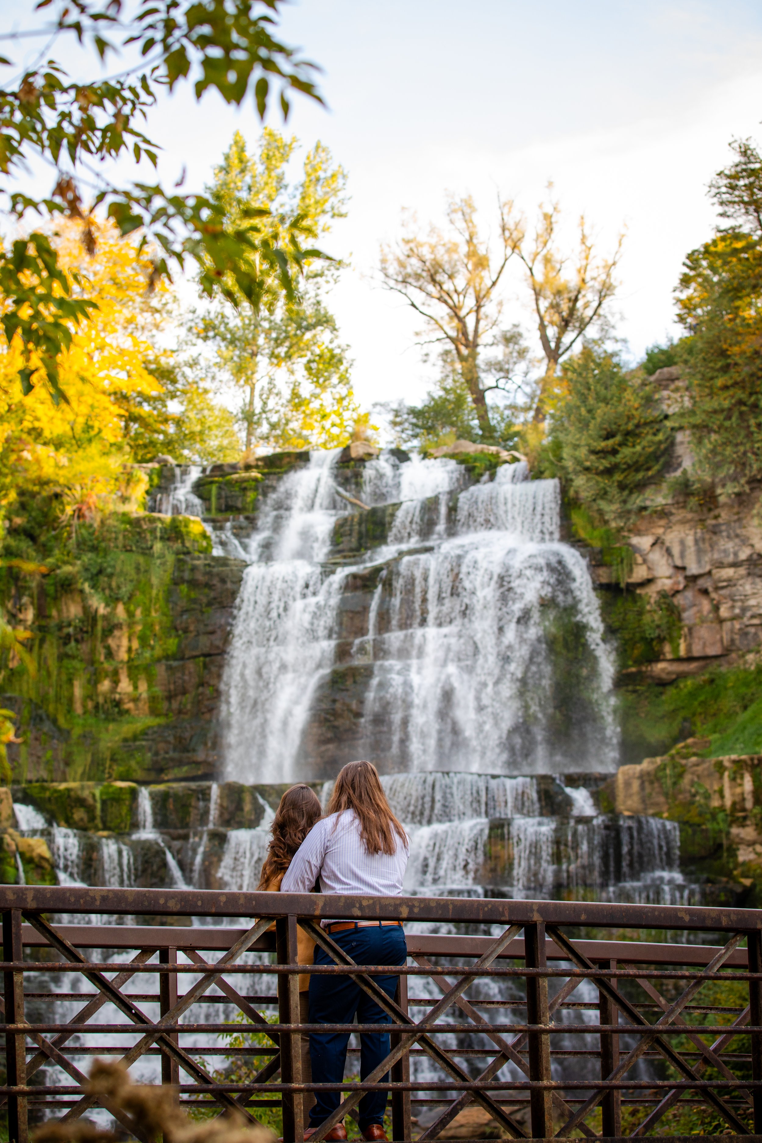 Two women standing on a wooden fence, looking at a large, multi-tiered waterfall surrounded by trees with green and autumn foliage.
